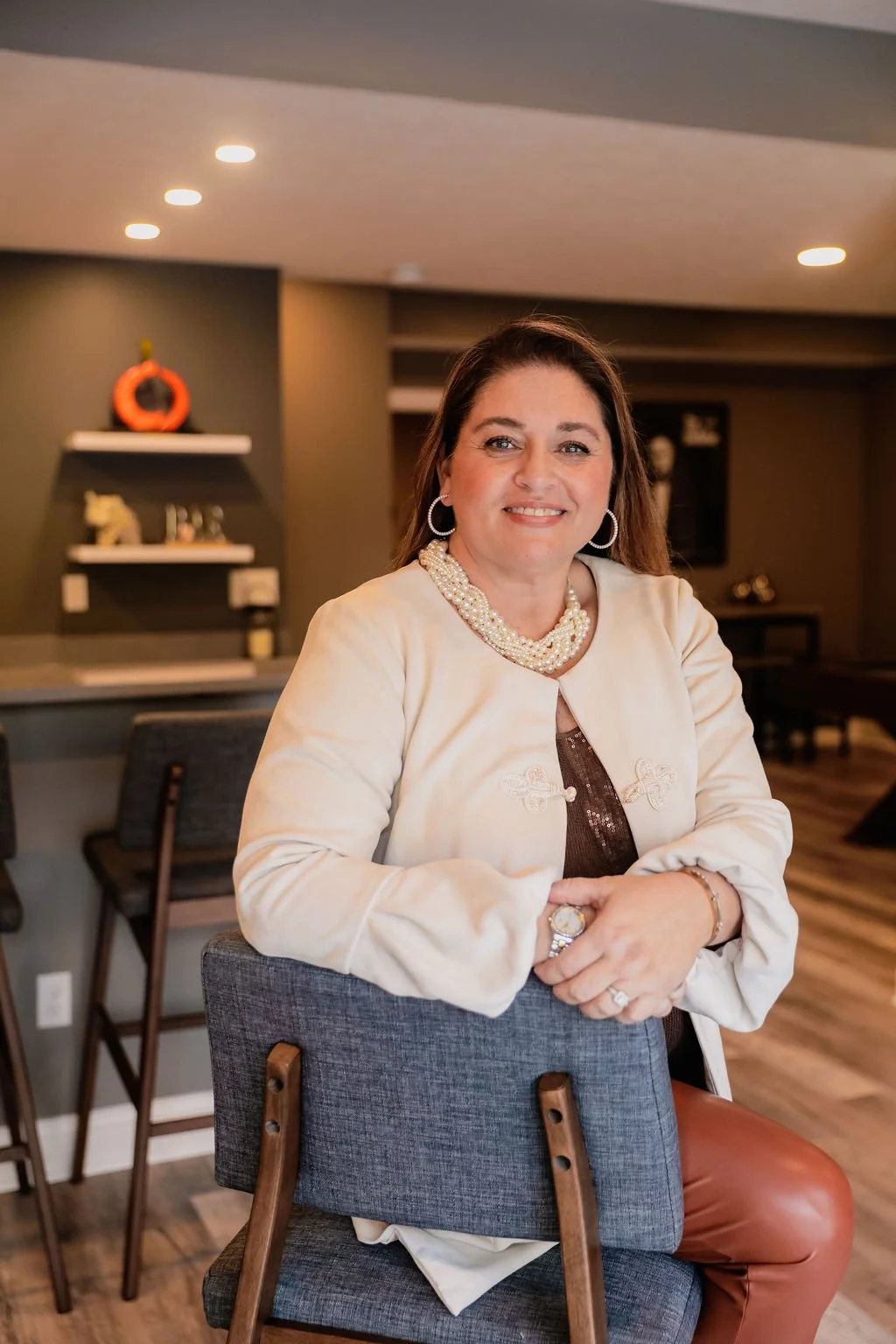 A woman with brown hair and hoop earrings smiling, sitting in a modern living room decorated with fall accents, including a pumpkin on a shelf behind her.