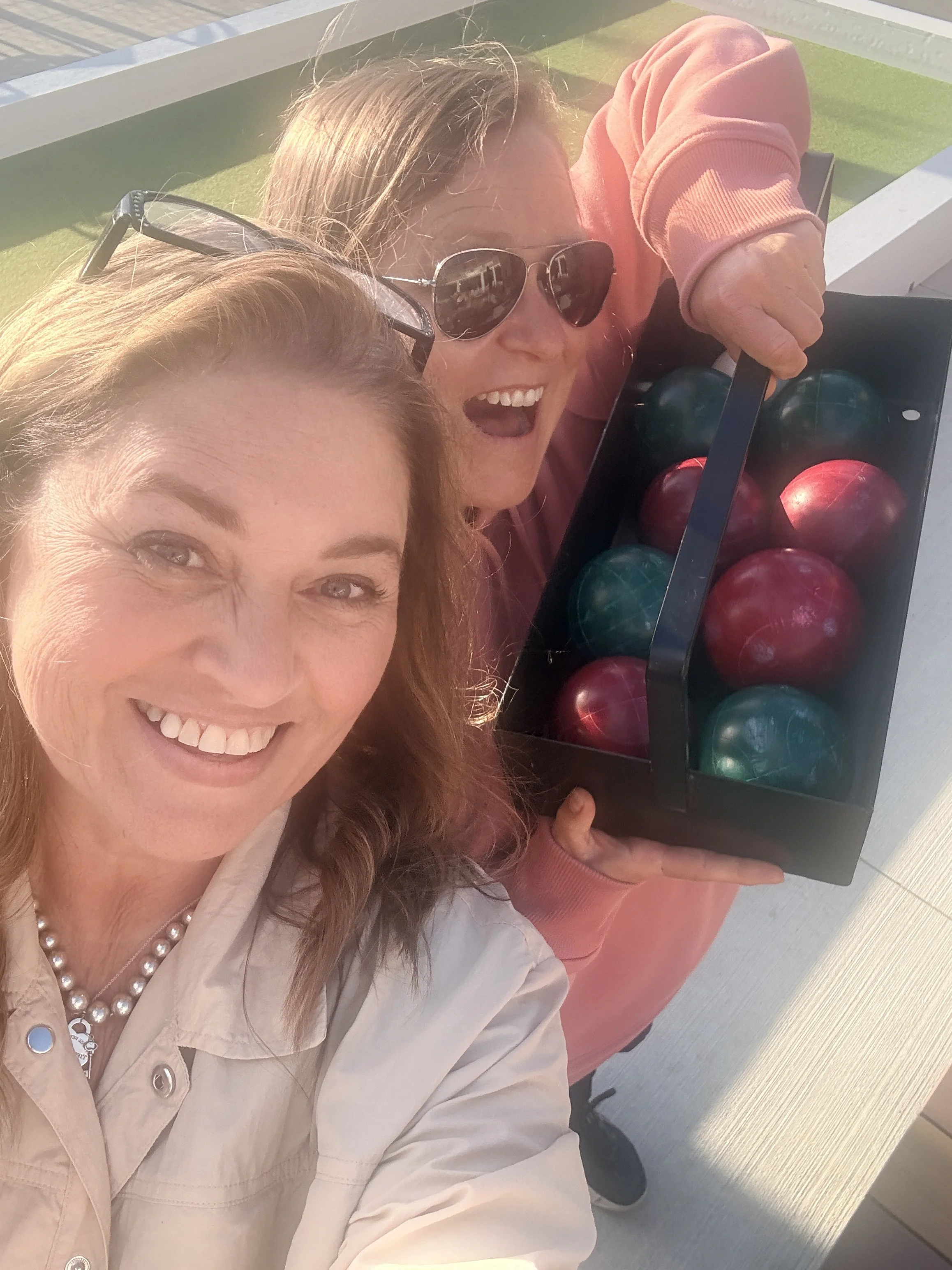 Two women taking a selfie, one of them holding a box of colorful Christmas ornaments, outdoors at a sunny location.
