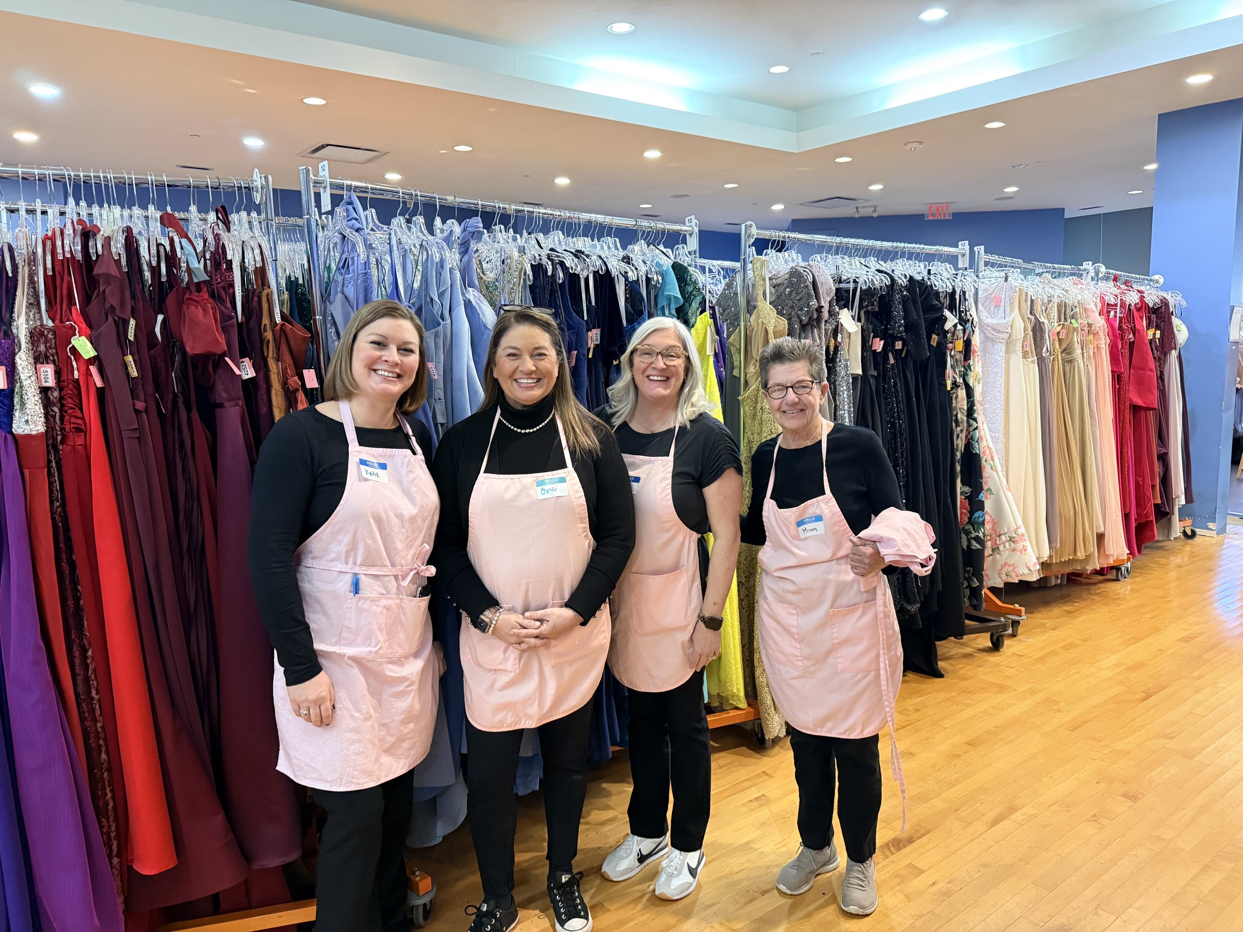 Four women standing in front of a clothing rack with colorful dresses at a store or boutique, smiling for the camera, wearing pink aprons and casual clothing.