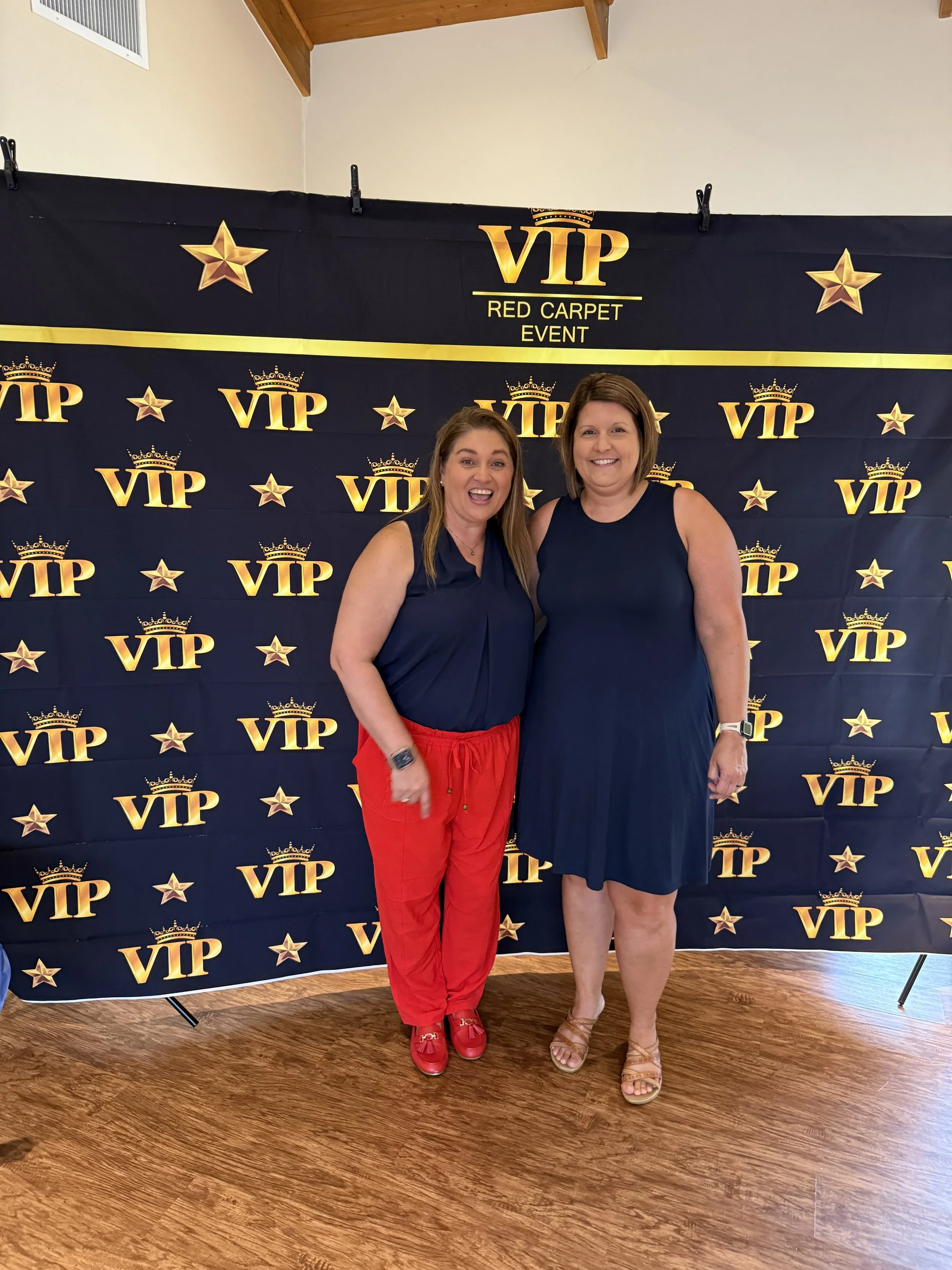 Two women standing in front of a VIP red carpet event backdrop, smiling and posing for the photo. The backdrop is decorated with gold stars and the VIP logo, with a wood floor and ceiling visible.
