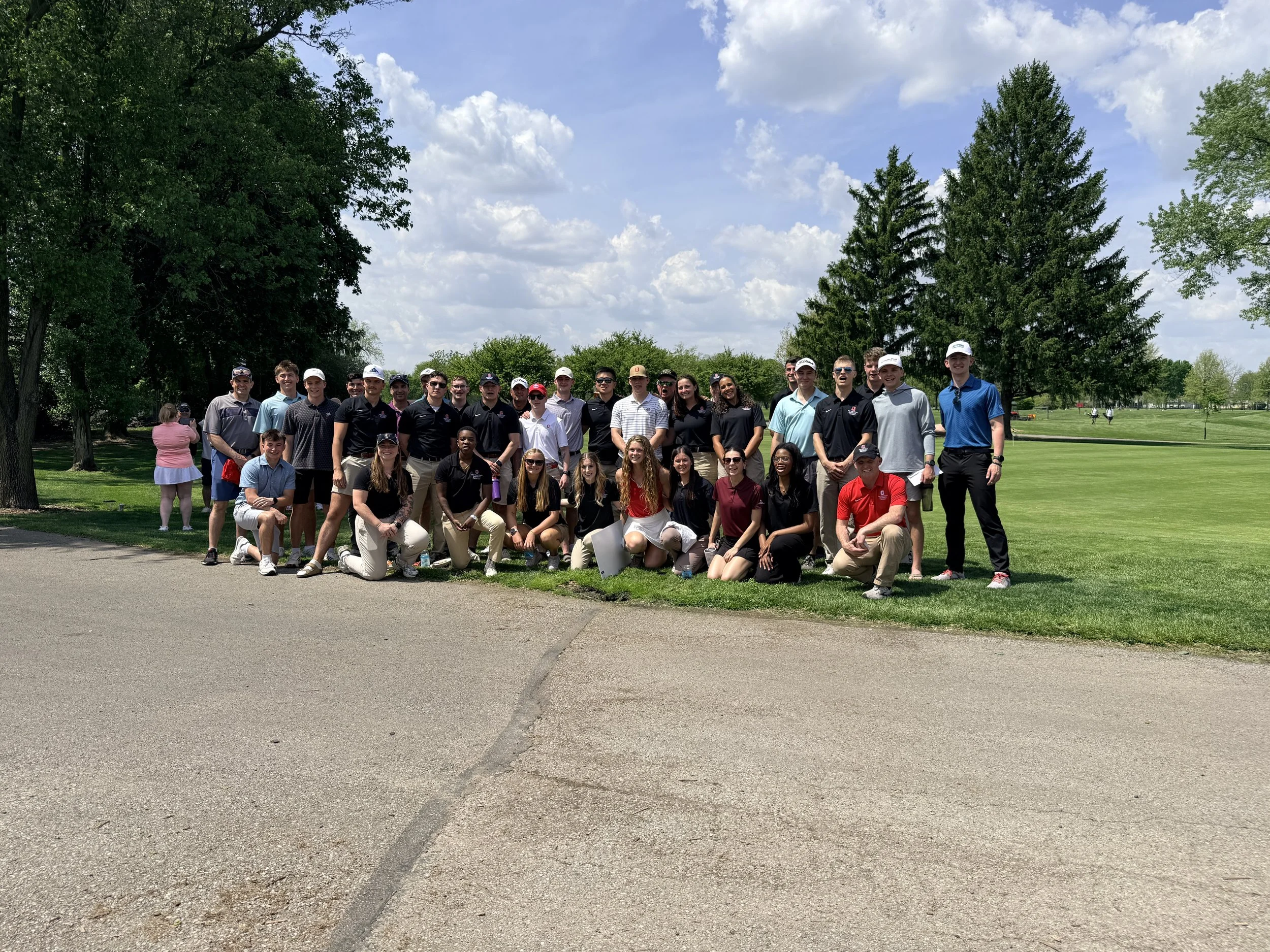 Group of young adults and teenagers posing for a photo on a golf course under a partly cloudy sky, with trees and greenery in the background.