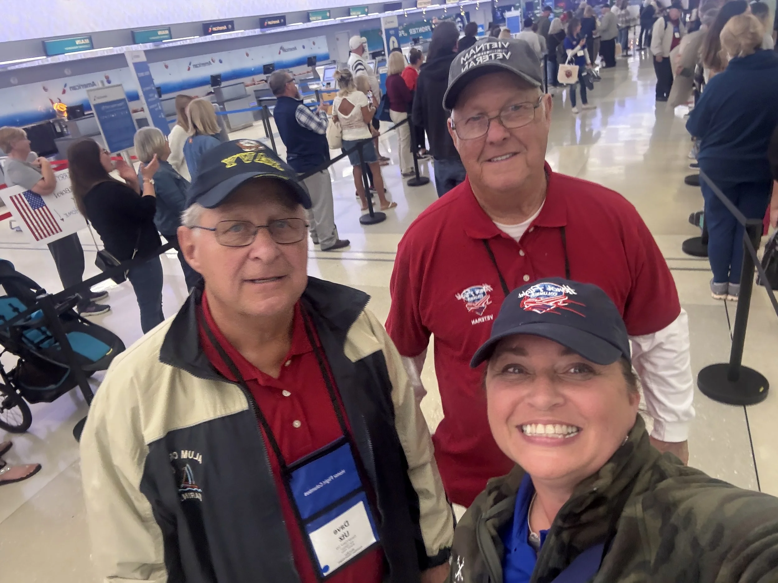 Smiling man in a red shirt and baseball cap taking a selfie with several people, crowded airport check-in area in the background.