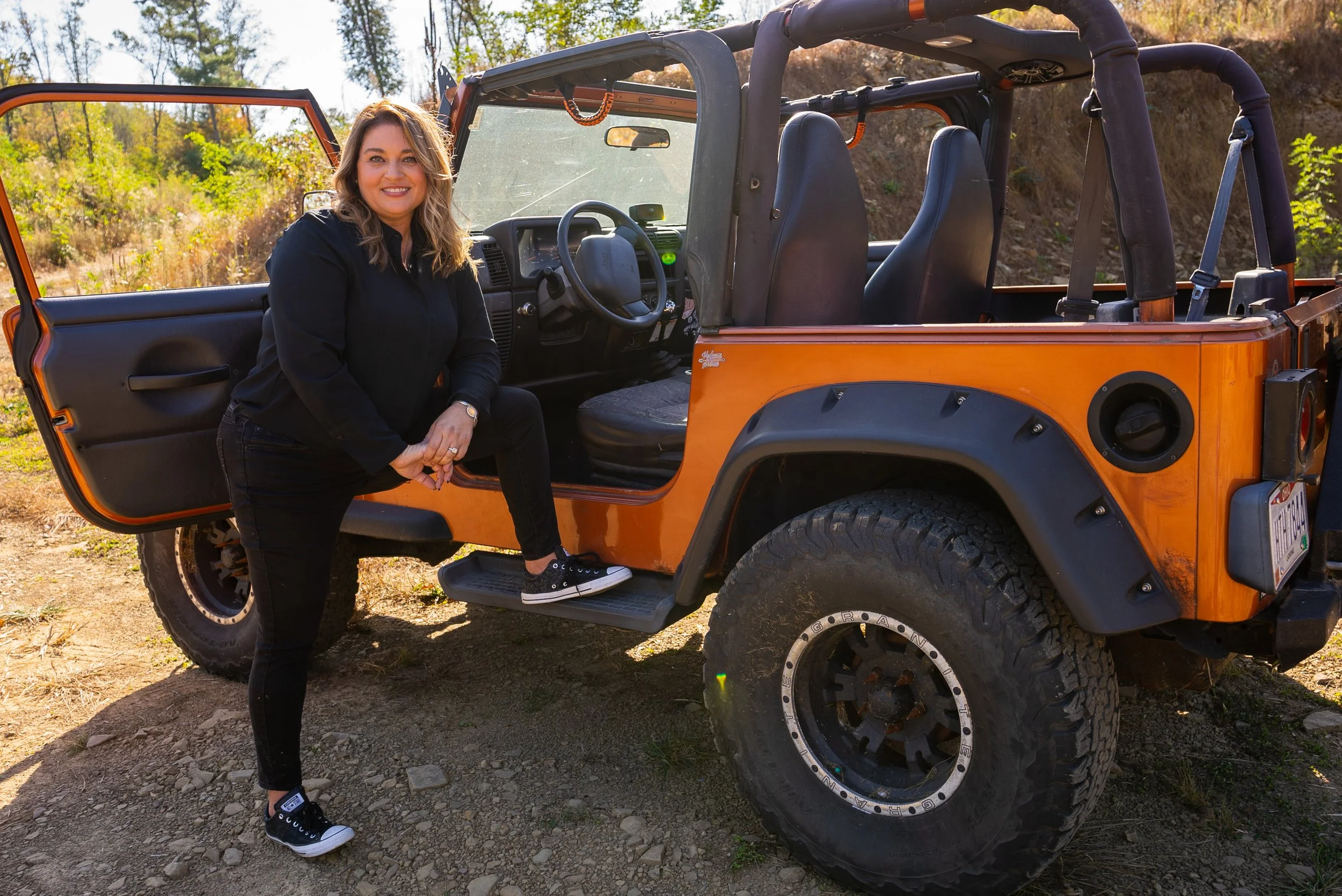 A woman in casual black attire and sneakers standing next to an orange off-road vehicle with a woman sitting inside, in a wooded outdoor setting with trees and sunlight.
