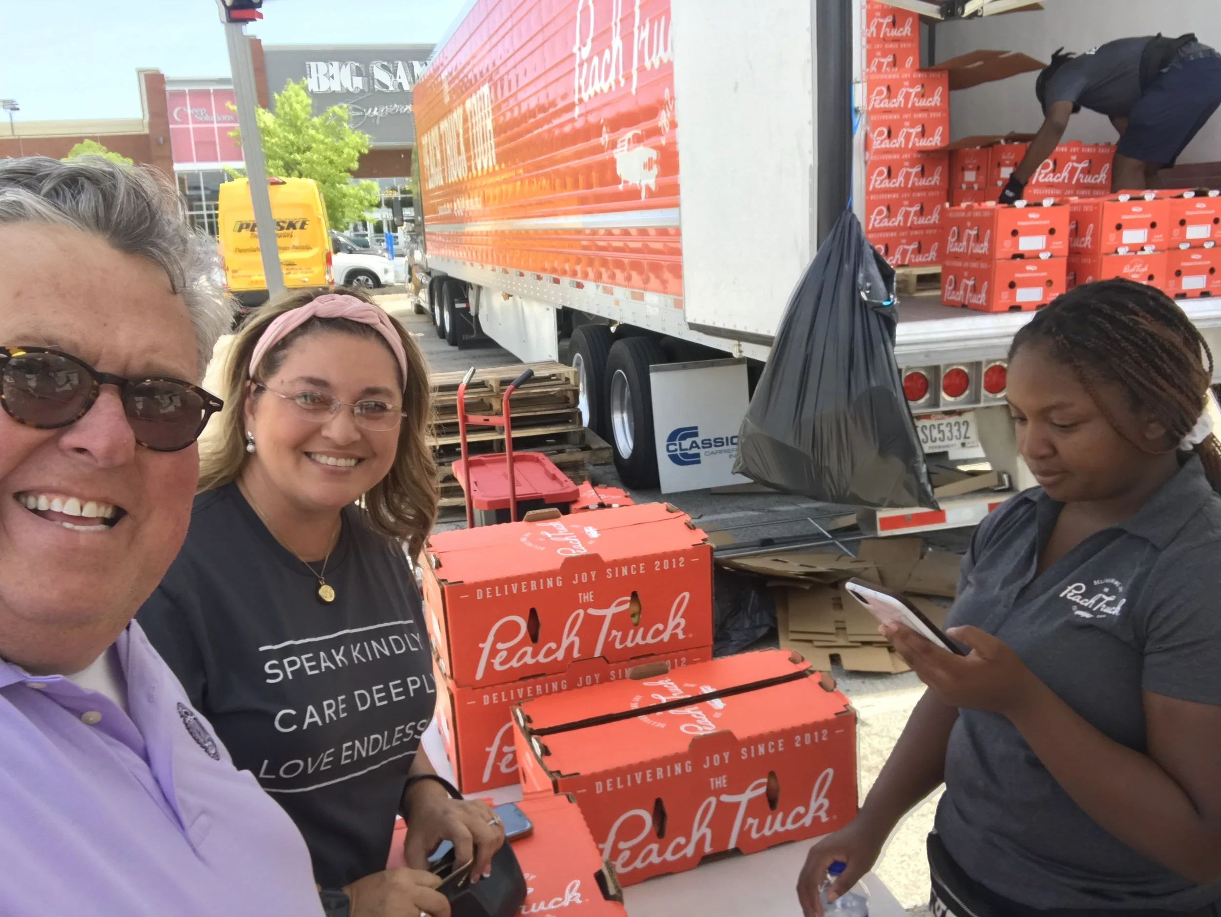 People standing near and organizing red Takeout boxes labeled 'Peach Truck' at an outdoor event with a large truck and parked cars in the background.
