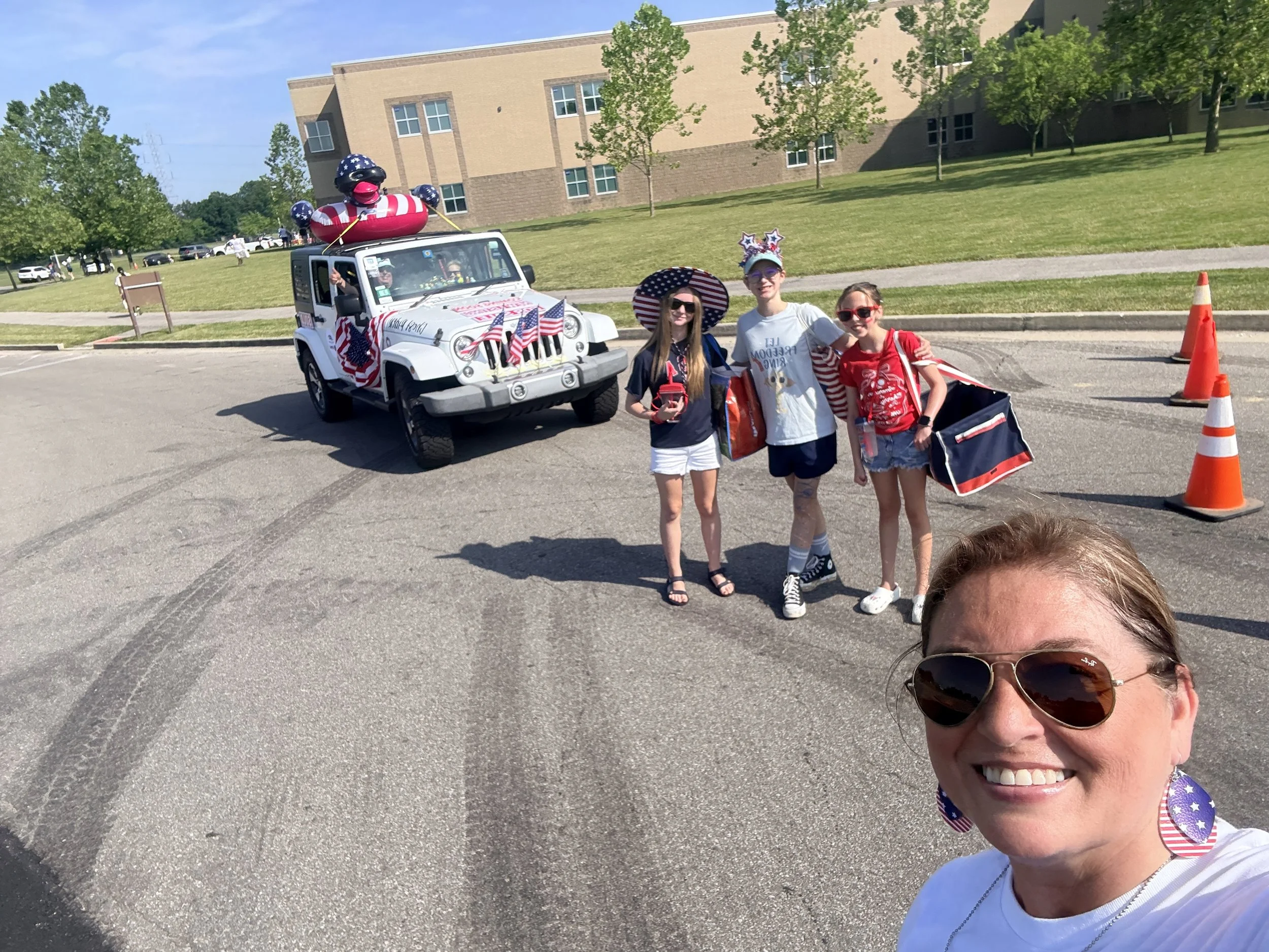 Group of four people standing in front of a decorated vehicle with American flags, large inflatable bear wearing a hat, and patriotic decorations, during a parade or celebration on a sunny day.