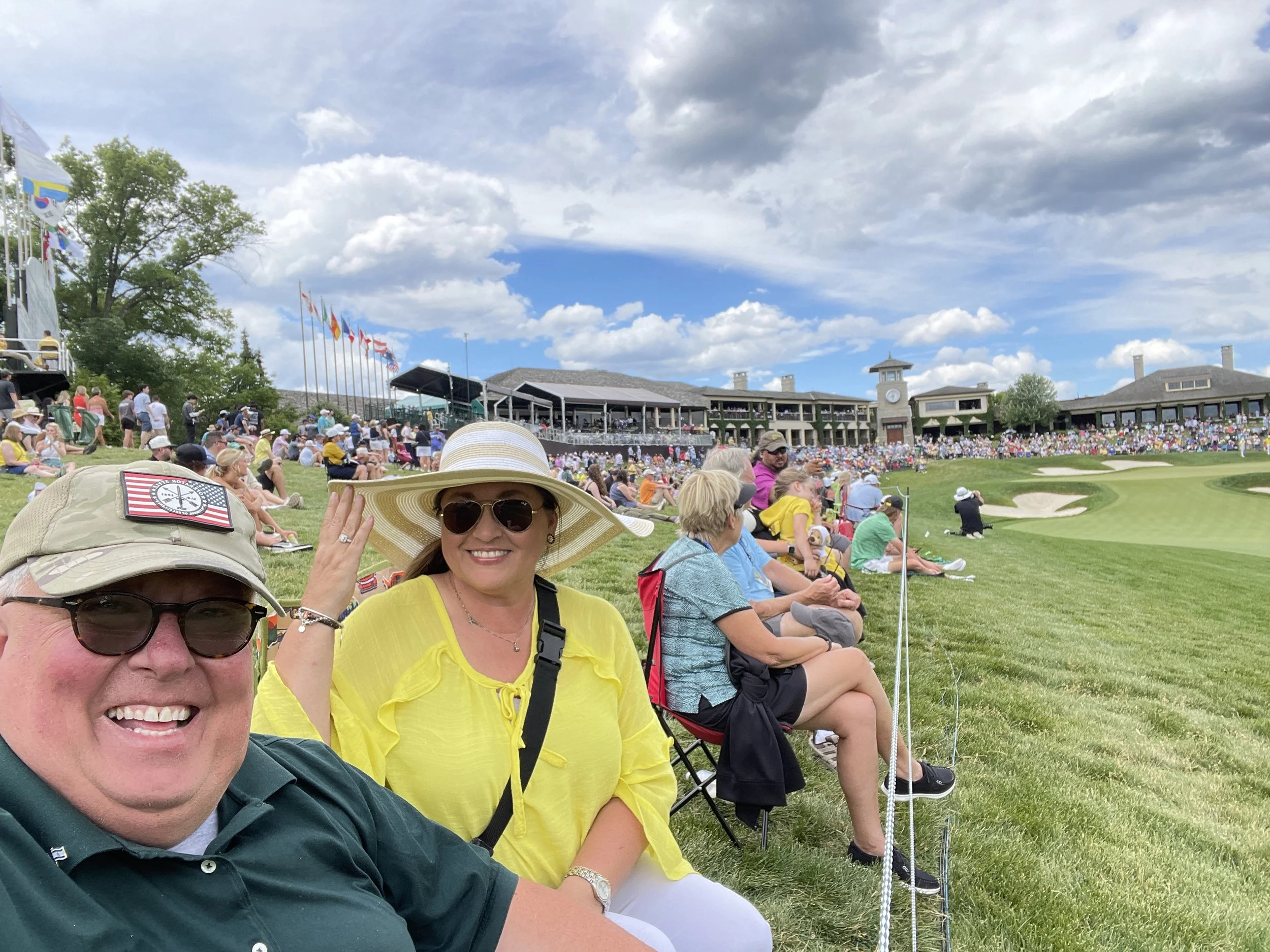 Smiling man and woman seated outdoors at a golf event, with many people in the background watching from a grassy area beside a golf course, beneath partly cloudy skies.