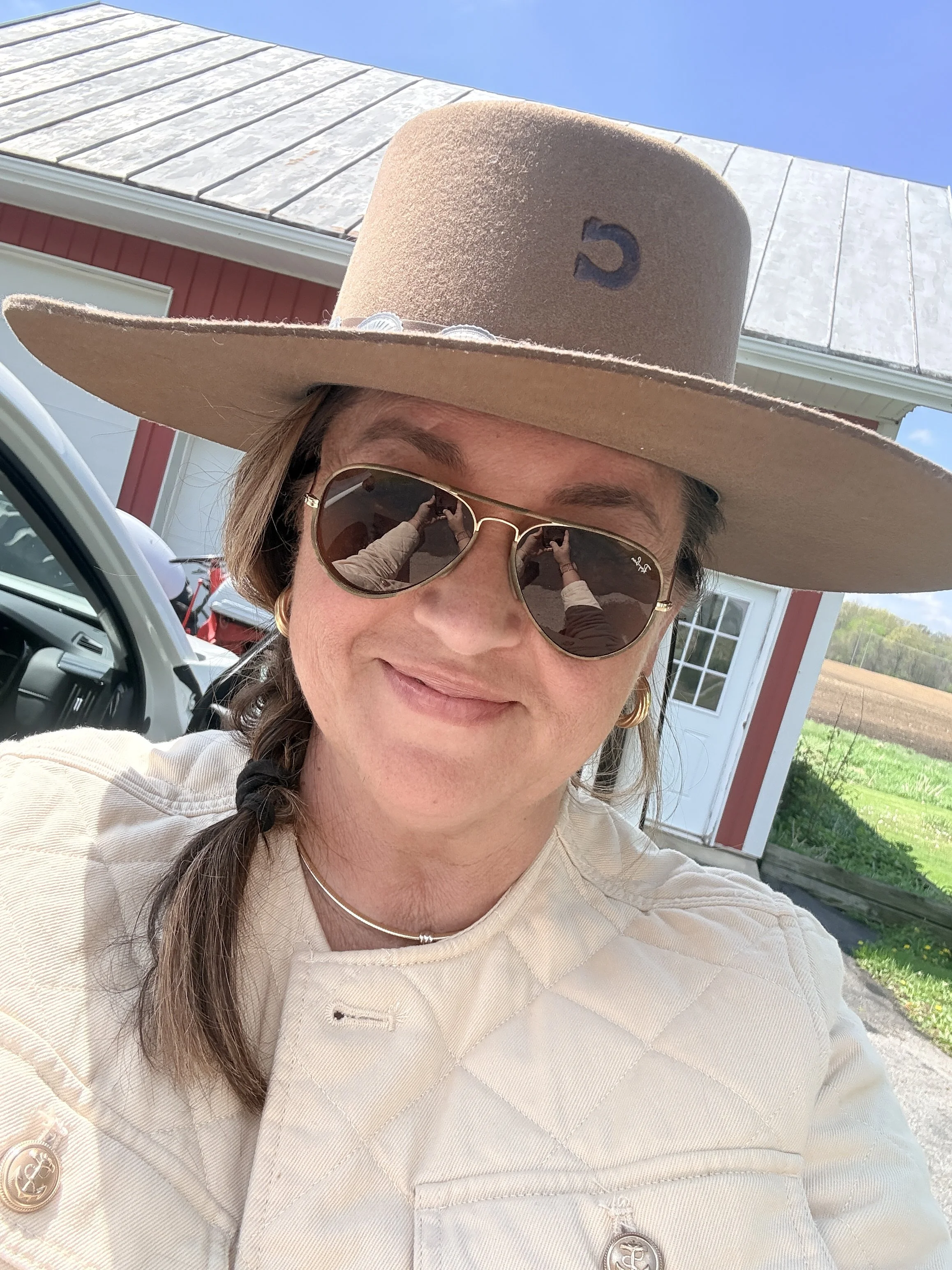 A woman taking a selfie outdoors, wearing a beige cowboy hat with a blue emblem, sunglasses, and a beige jacket, standing in front of a building with a red and white exterior, with cars and a grassy area visible in the background.