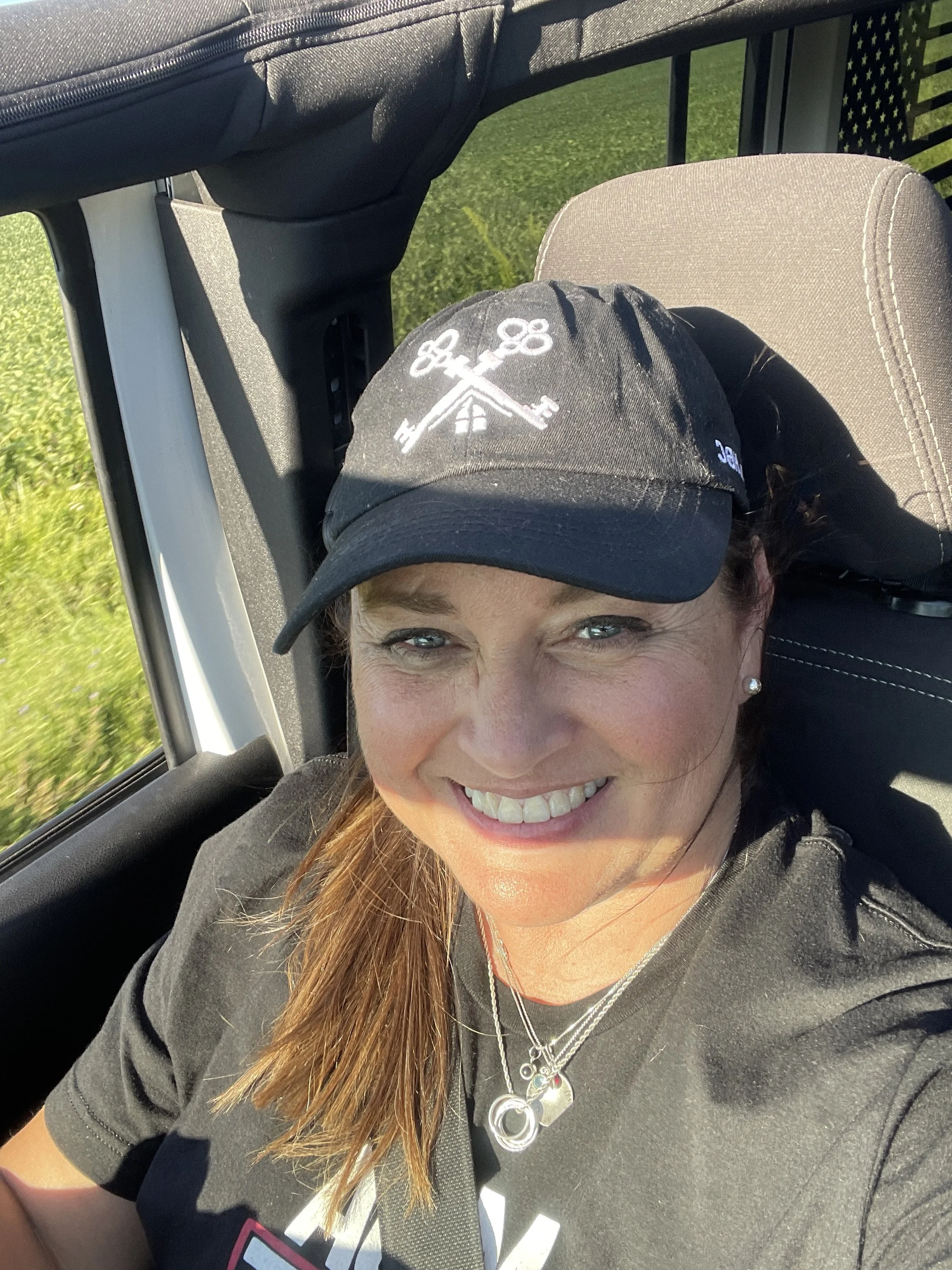 Close-up of a smiling woman with brown hair wearing a baseball cap, sitting in a car with green grass outside the window.