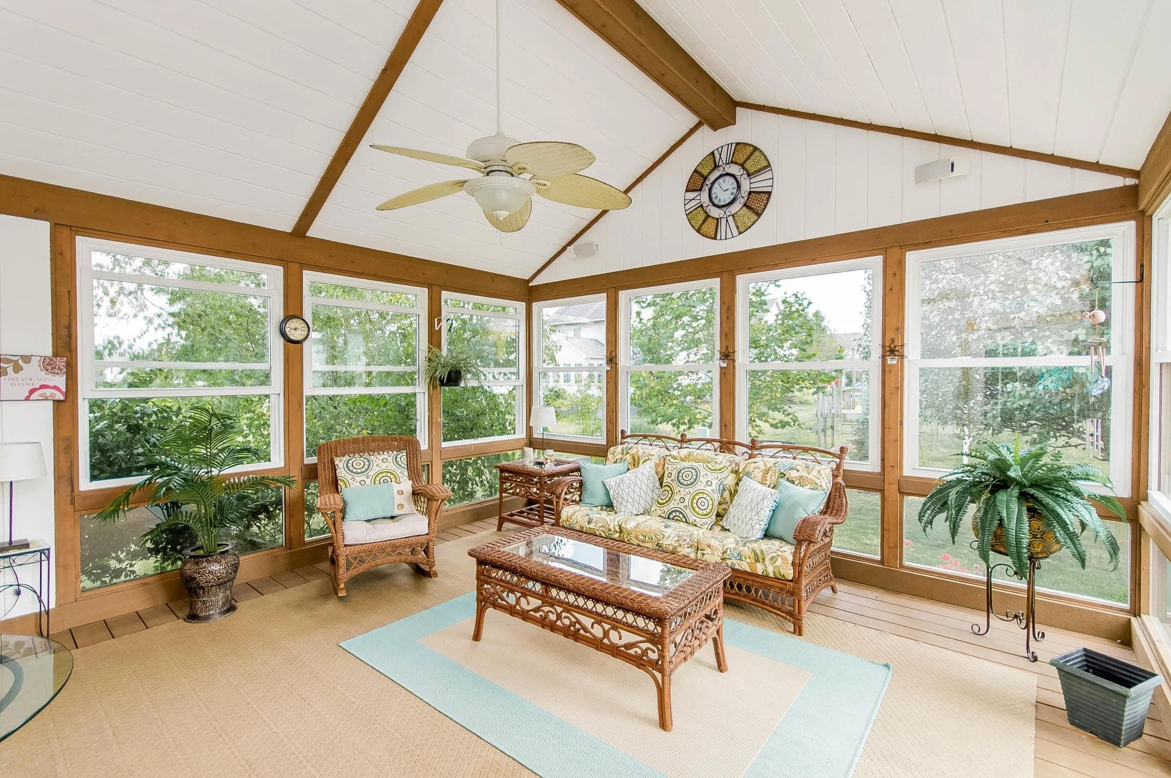 Sunroom with wicker furniture, large windows, ceiling fan, and clocks on the walls, with plants and a view of a backyard outside.