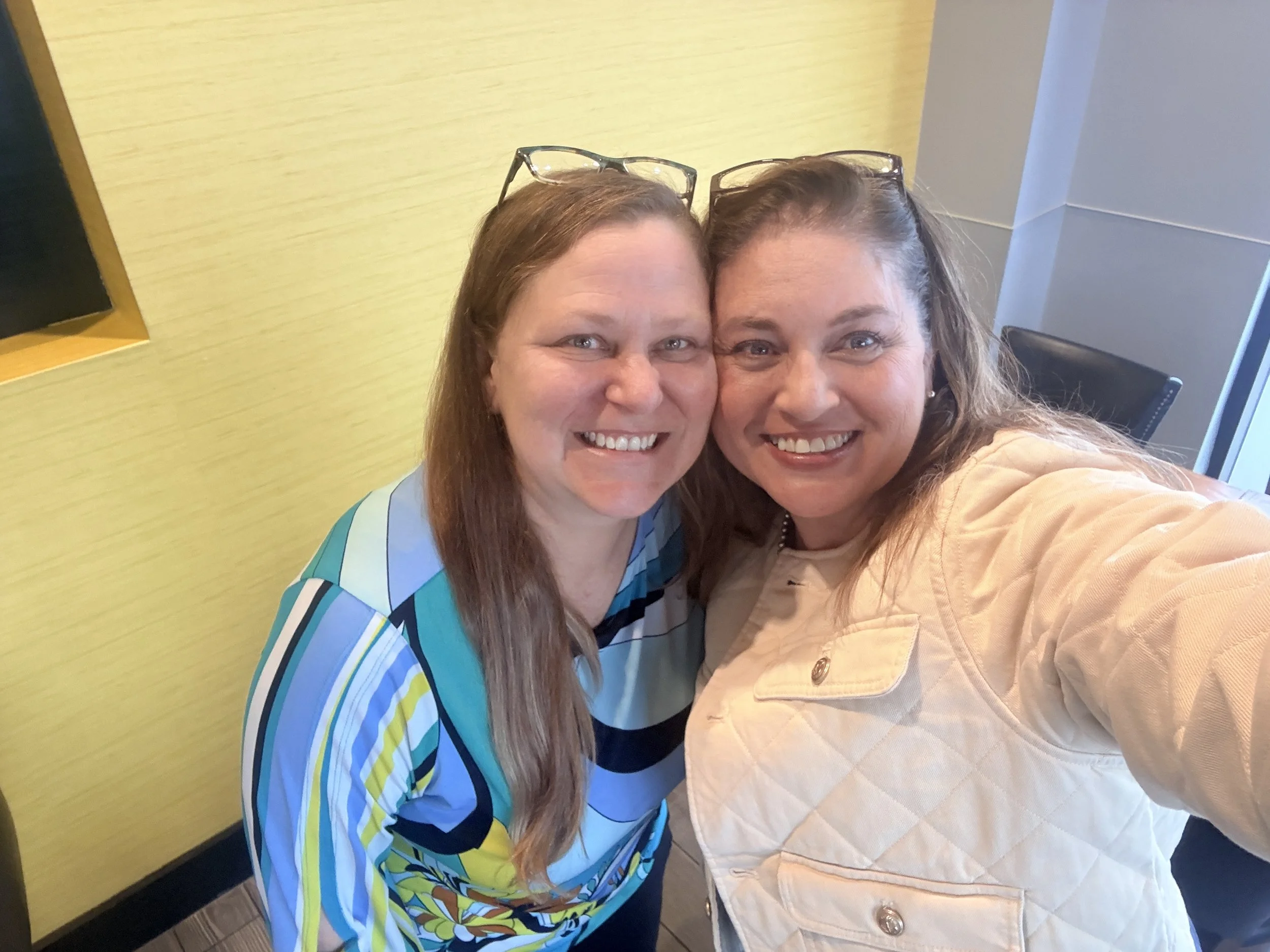 Two women smiling and taking a selfie together indoors near a yellow wall and a window, both wearing glasses on their heads.