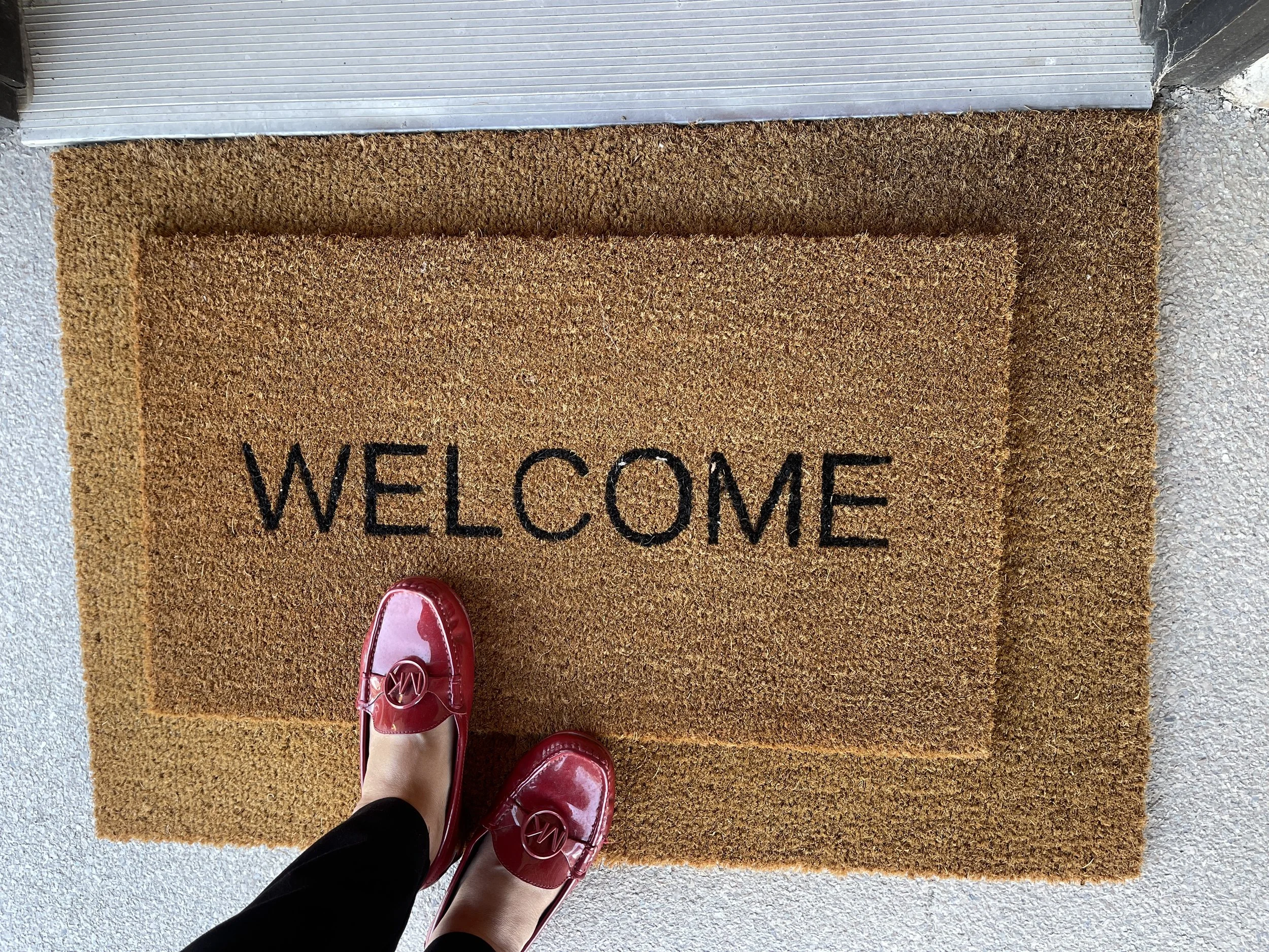 A front door mat reading "WELCOME" with two people wearing pink shoes standing on it, taken from above.