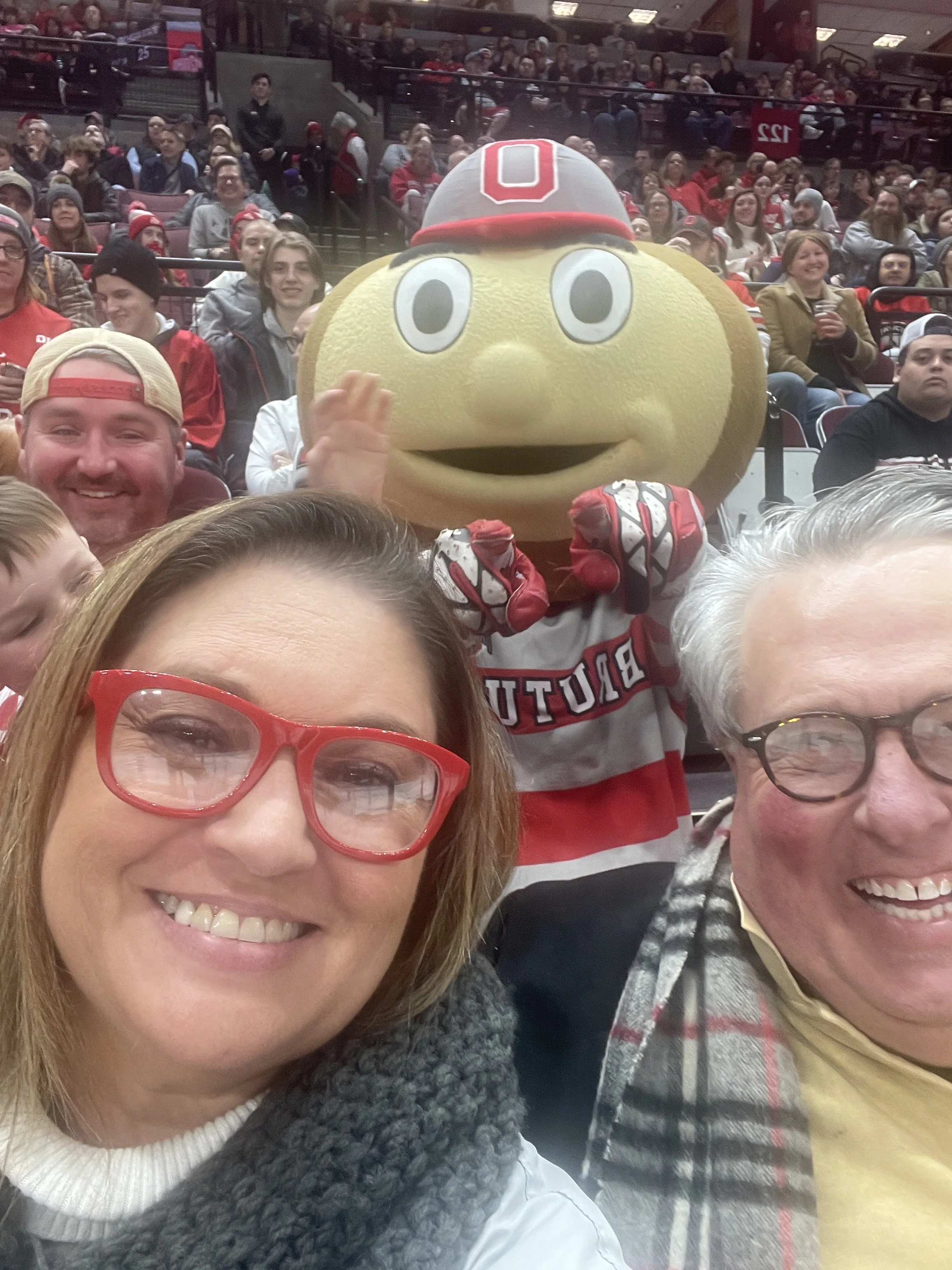 People at a sporting event, with one person wearing a large mascot costume resembling a yellow piece of bread with big eyes and a red football helmet, in the stands filled with spectators.