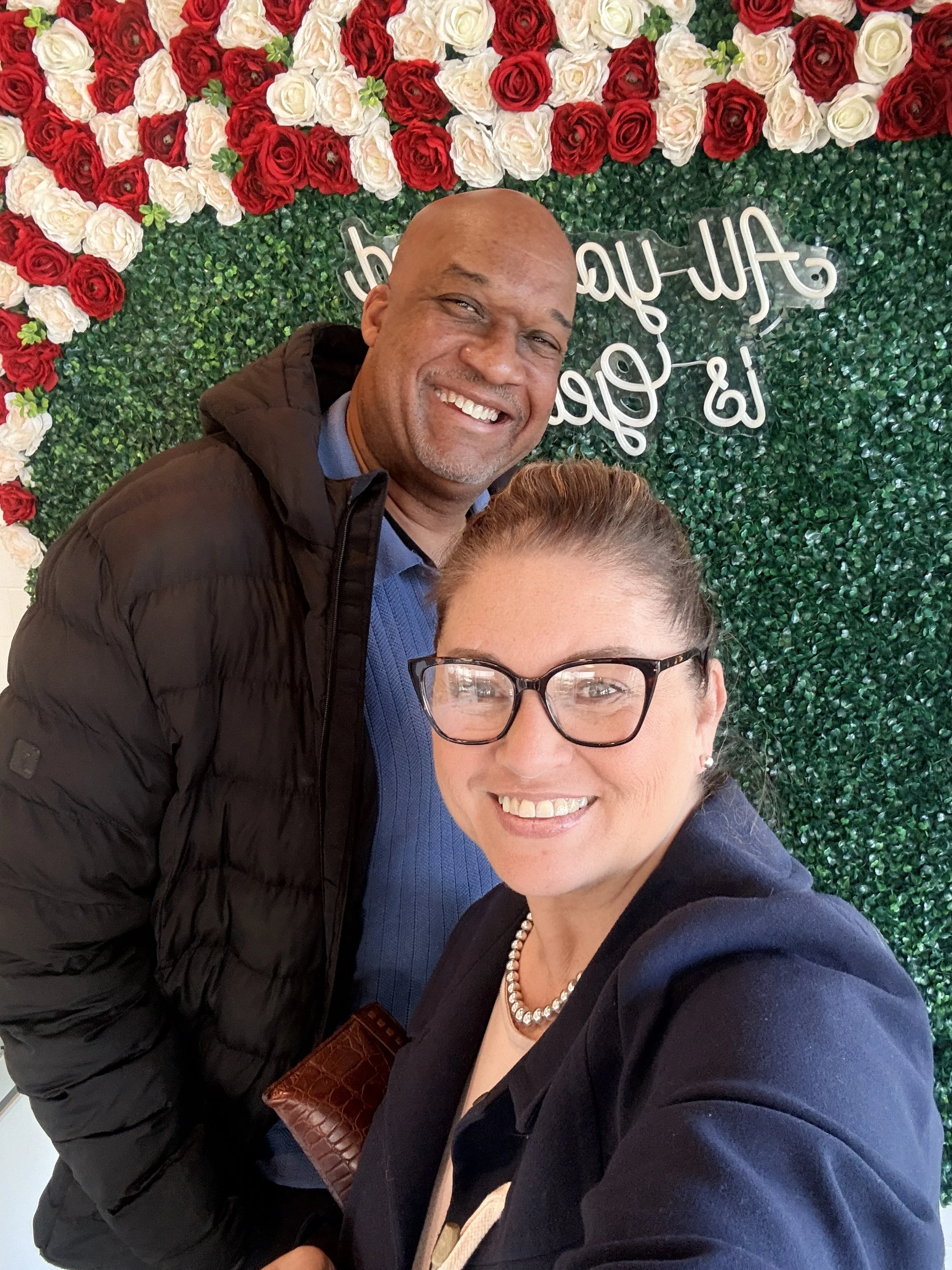 Two people taking a selfie in front of a wall decorated with red and white roses and greenery, with a neon sign.