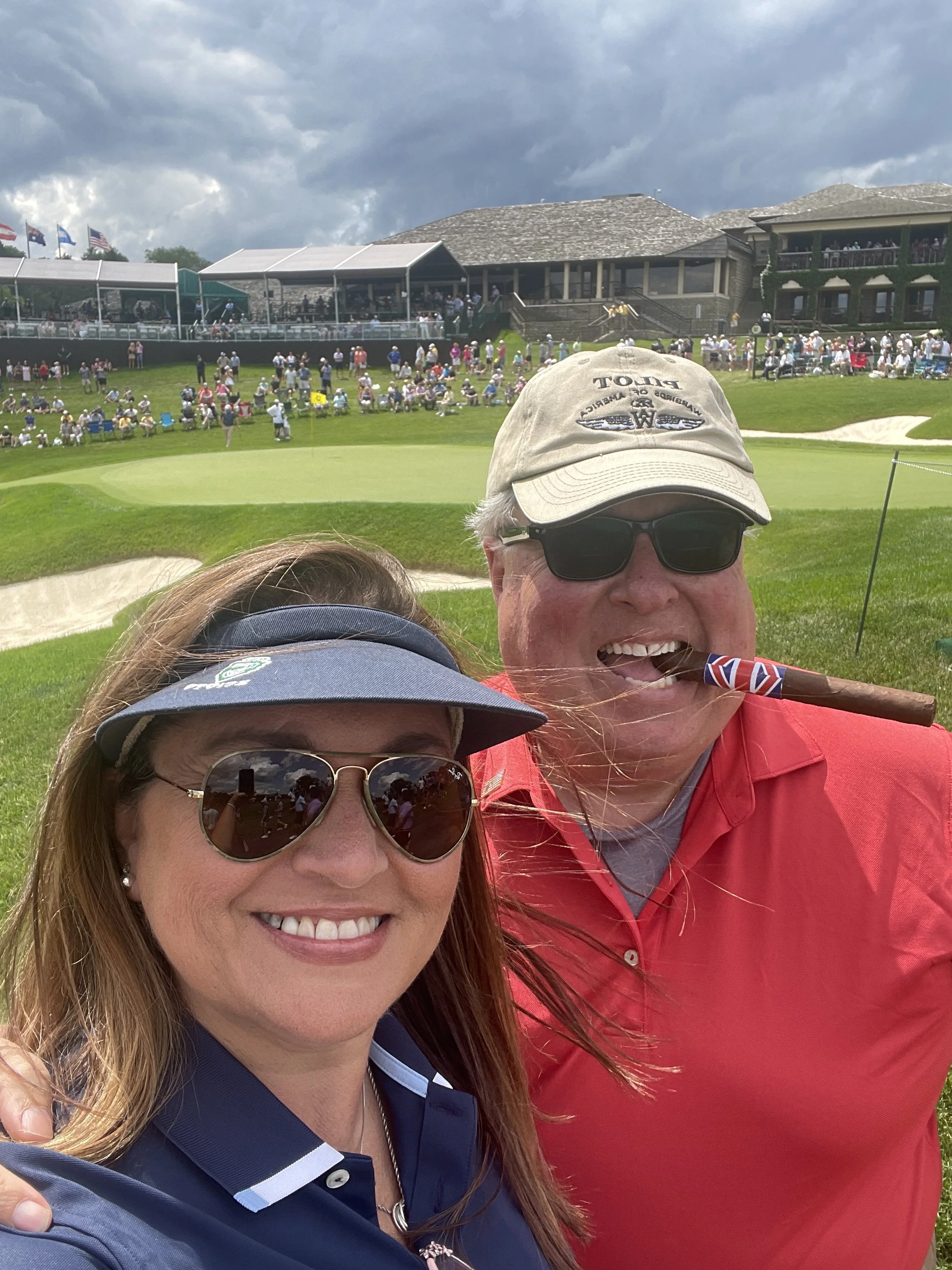 Two smiling people taking a selfie with a golf course and a clubhouse in the background. The man is wearing a beige golf cap, sunglasses, a red shirt, and is holding a cigar with a Union Jack band. The woman is wearing sunglasses, a navy blue golf sh