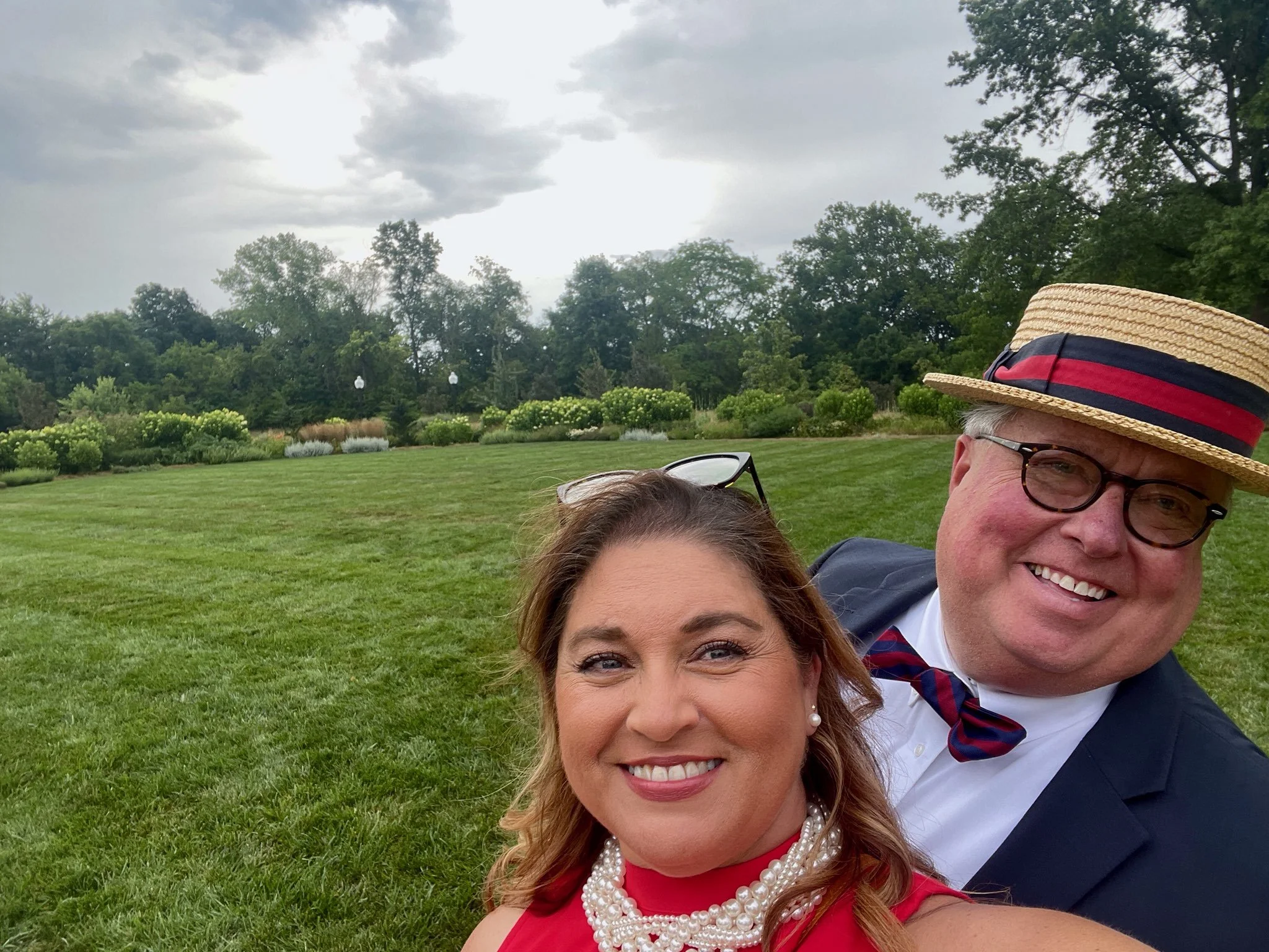 Close-up of smiling woman and man in formal attire with hat, outdoors on cloudy day, with lush green park, trees, and cloudy sky in background.