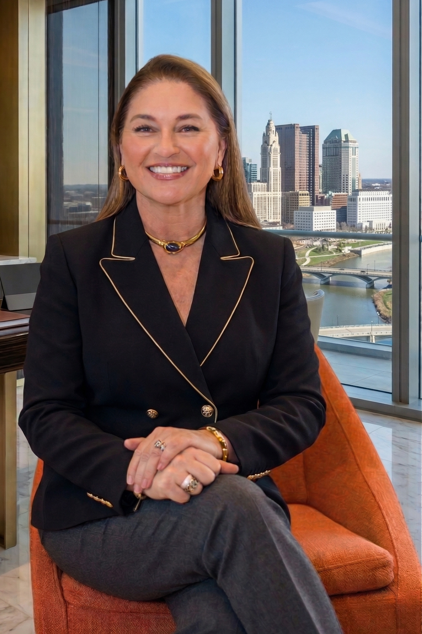 A smiling woman with shoulder-length brown hair, wearing gold jewelry and a black blazer, sitting on an orange chair in front of large windows with a city skyline in the background.