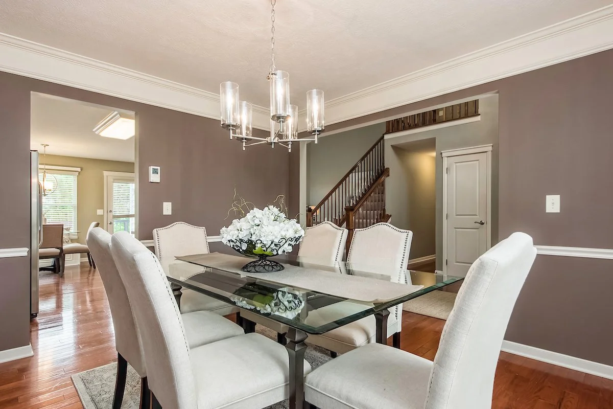 A dining room with a glass-top table, six white upholstered chairs, a flower centerpiece, a modern chandelier, and hardwood floors, with a staircase and a doorway leading to another room in the background.