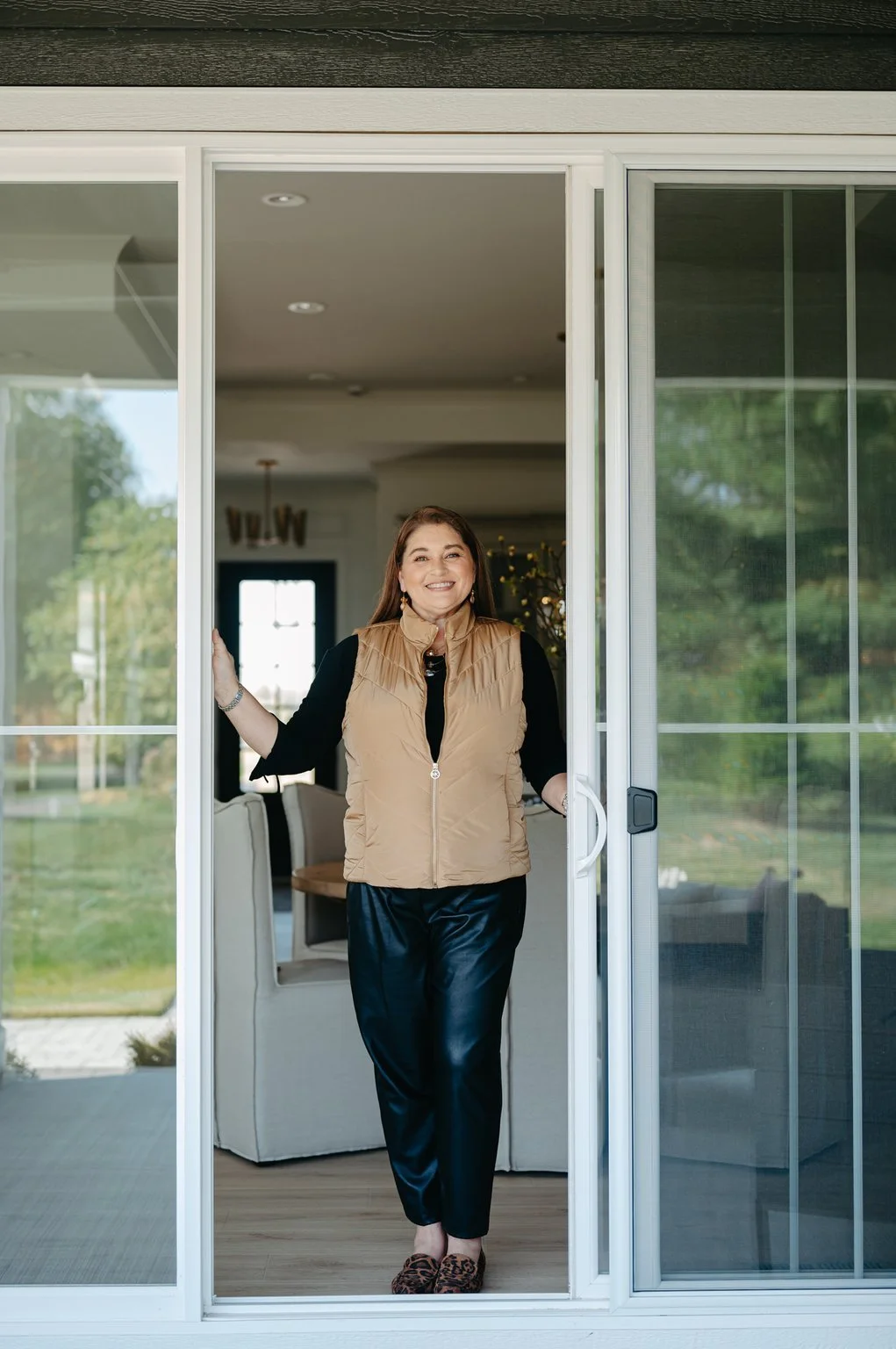 A woman standing at a sliding glass door, smiling inside her home with a garden visible outside.