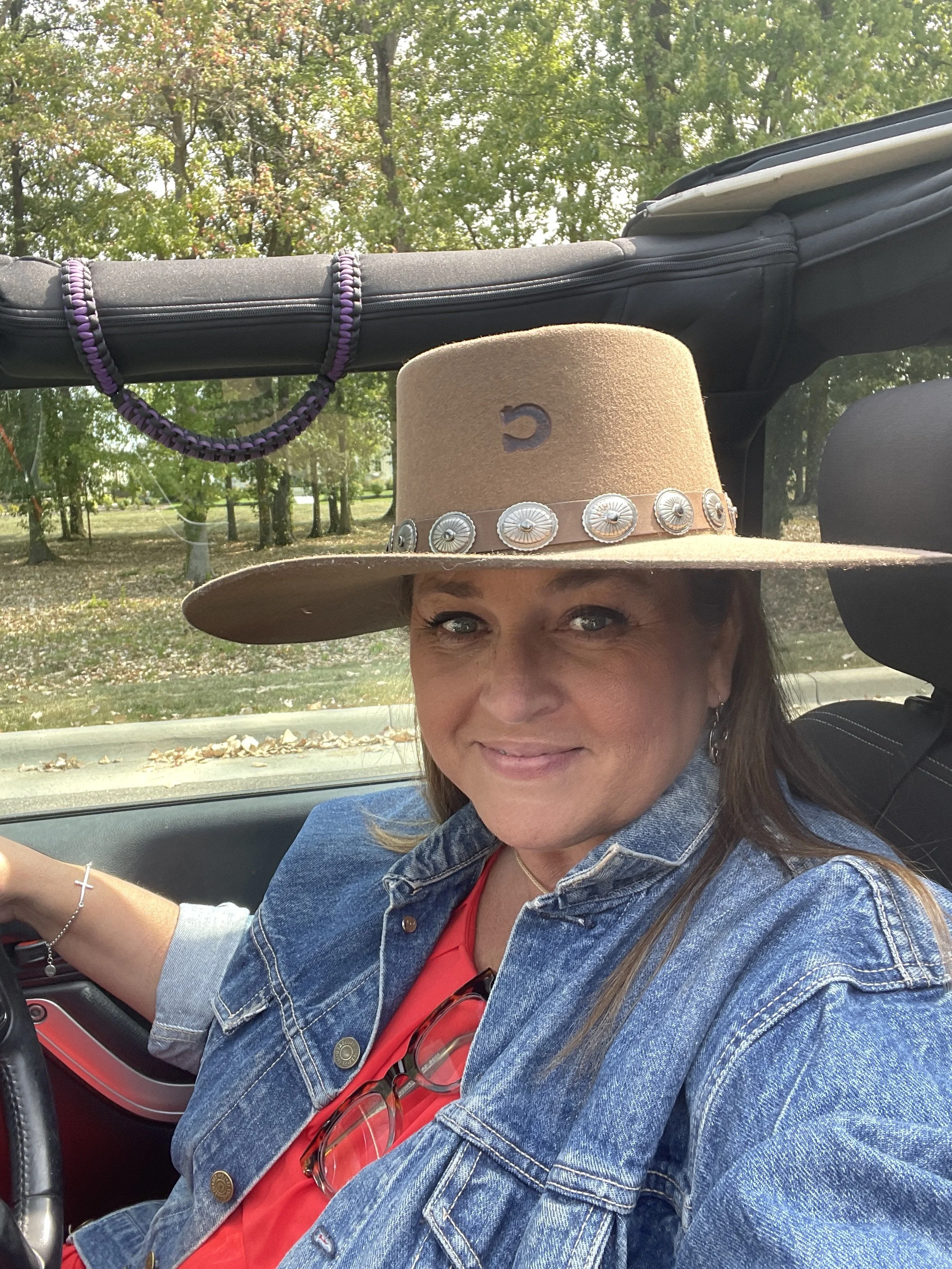 A woman with long hair and light skin, wearing a denim jacket and red shirt, smiling while sitting in a car, with a large cowboy hat decorated with silver conchos on her head, showing only her lower face and eyes. The car interior is visible, with gl