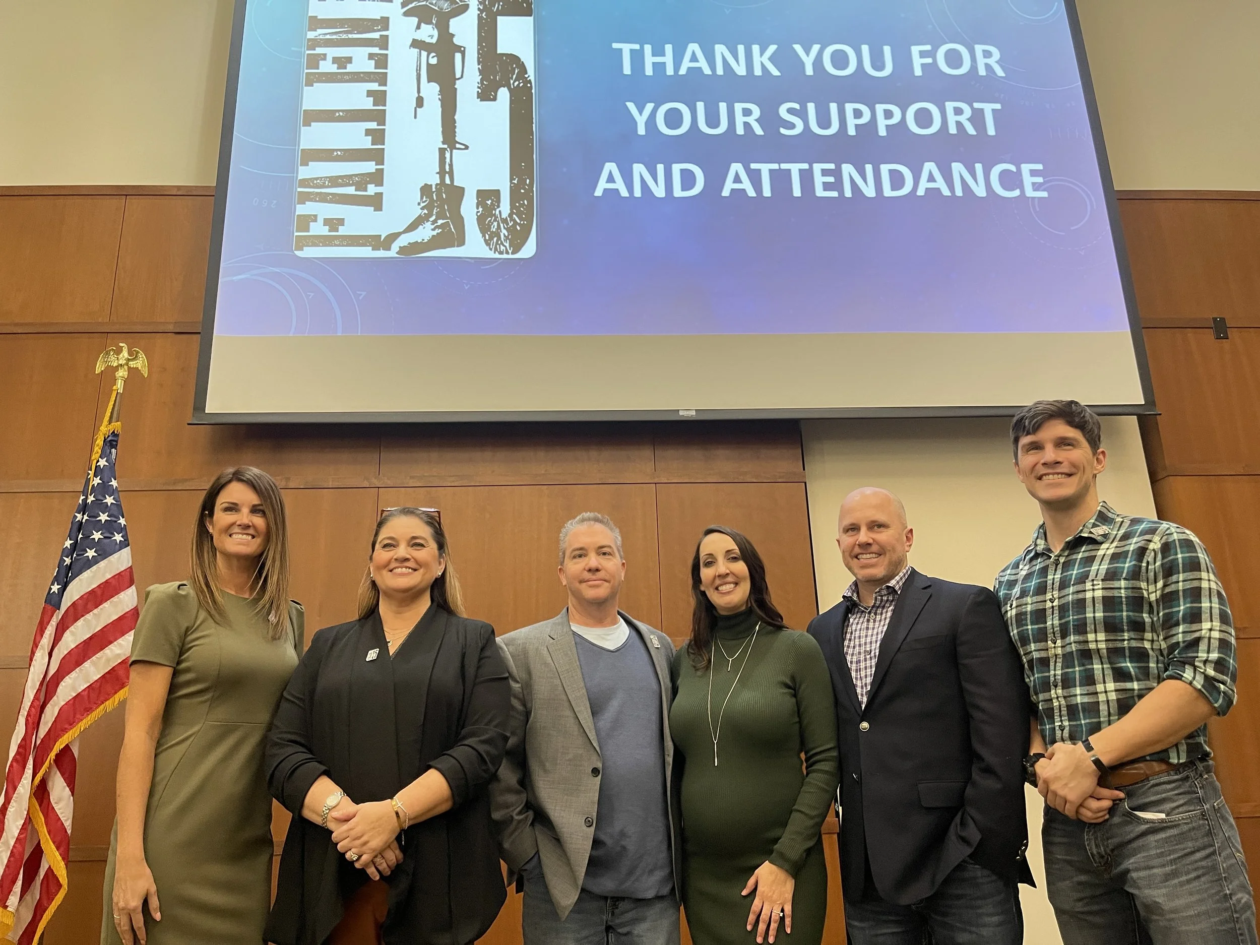 A group of six people posing for a photo in front of a presentation screen that reads "Thank you for your support and attendance." The group includes four women and two men, all smiling. An American flag and a military flag are visible on the left si