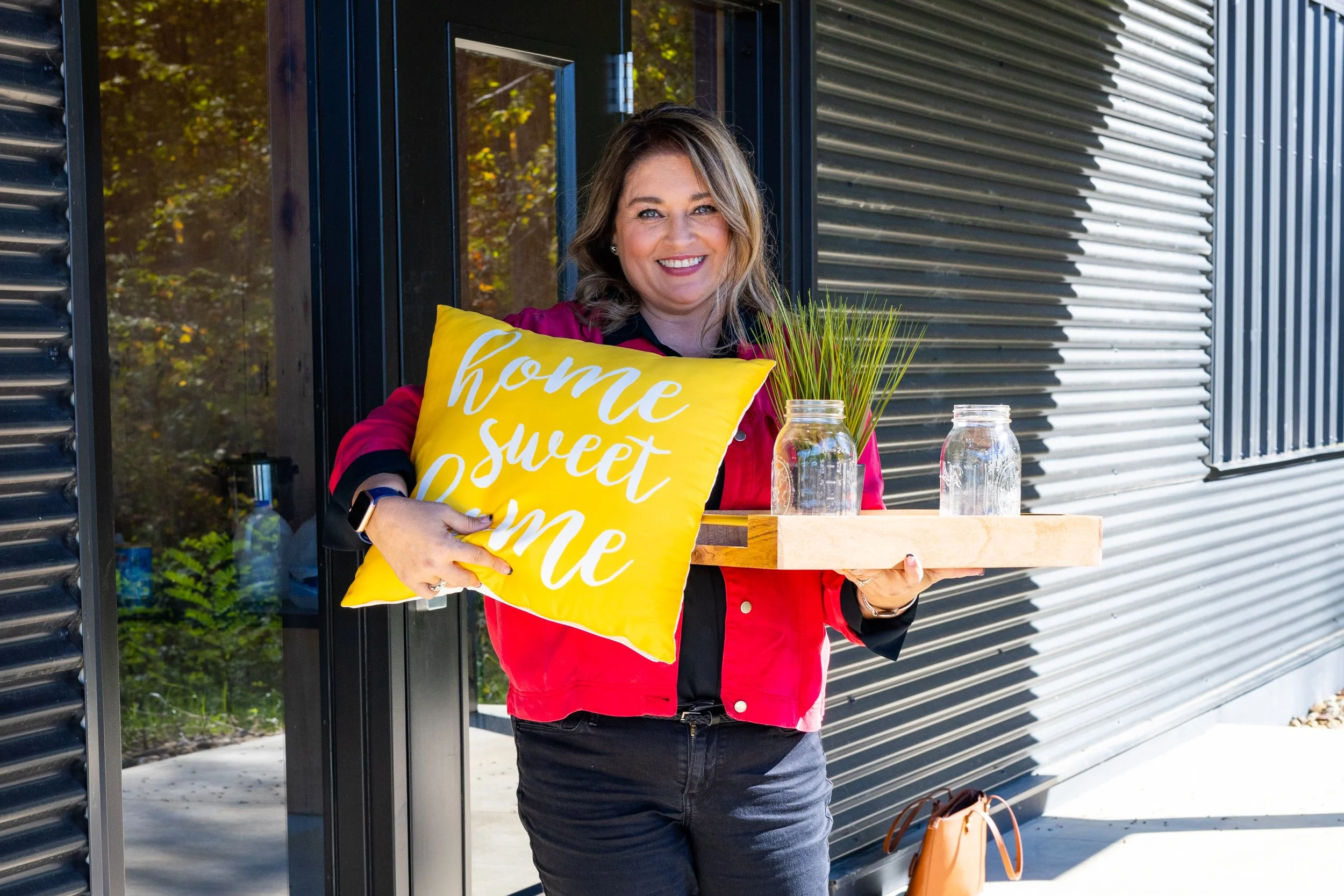 A woman with wavy, shoulder-length hair holds a yellow pillow with white text that says 'home sweet home' and a wooden tray with two glass jars and some green grass, standing outside a modern building with dark metal siding.
