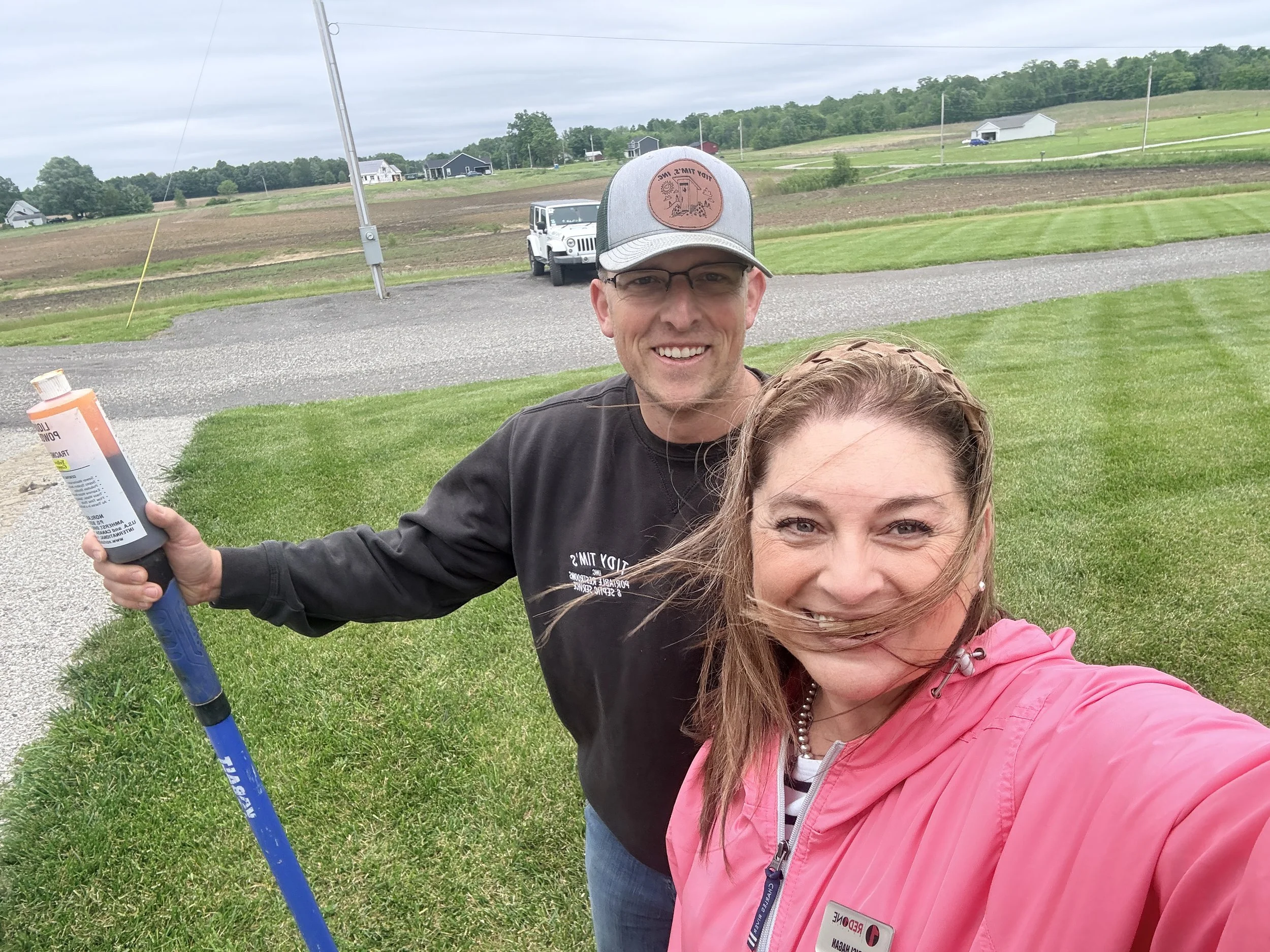 A smiling man in a gray cap and black hoodie standing outside on a lawn, holding a pole with a bottle attached. A woman in a pink jacket is partially visible in the foreground. In the background, there is a rural landscape with fields, houses, and a 