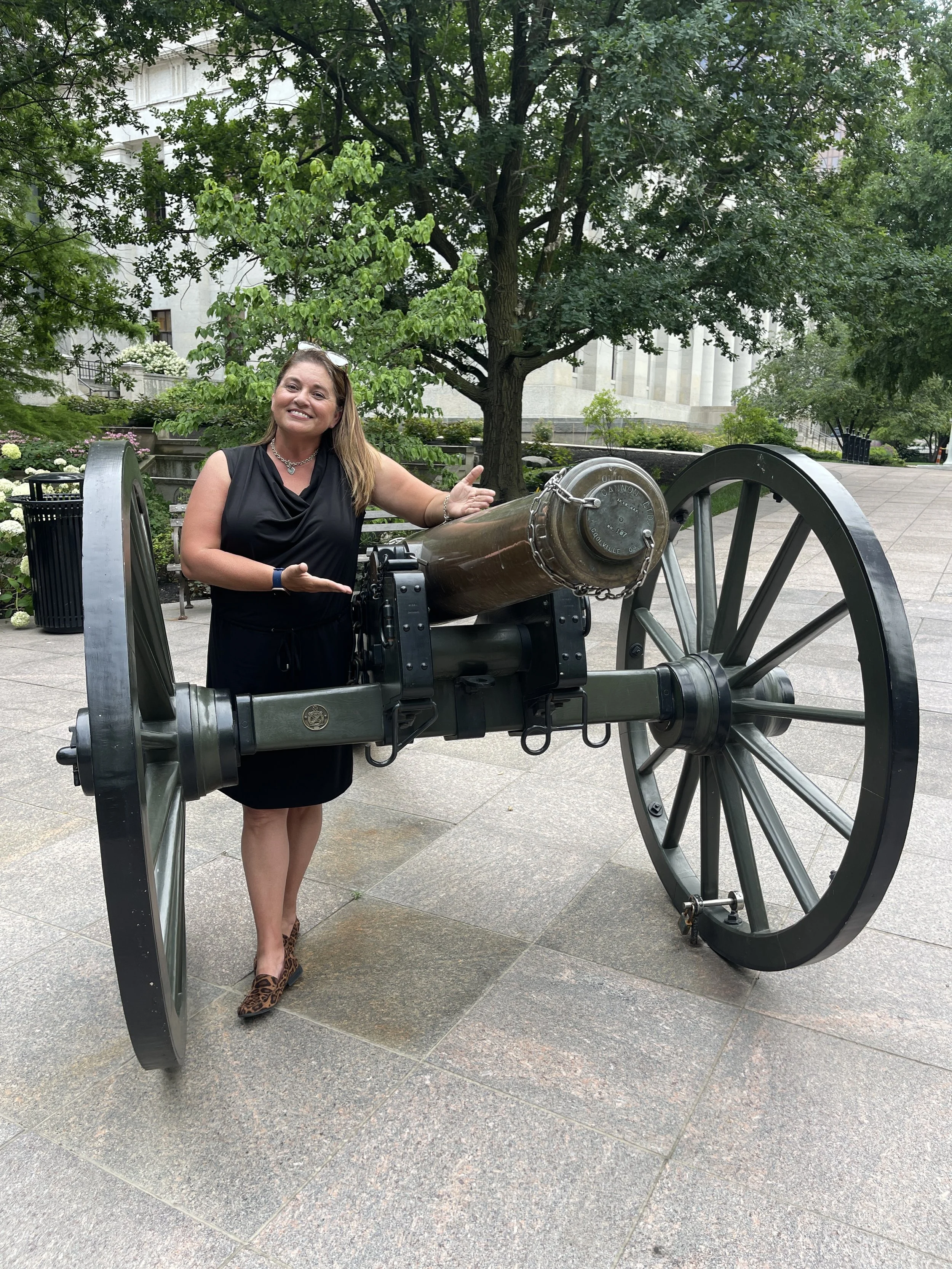 A woman in a black dress and leopard print heels standing next to an old-fashioned cannon outdoors in a park, with green trees and a white building in the background.