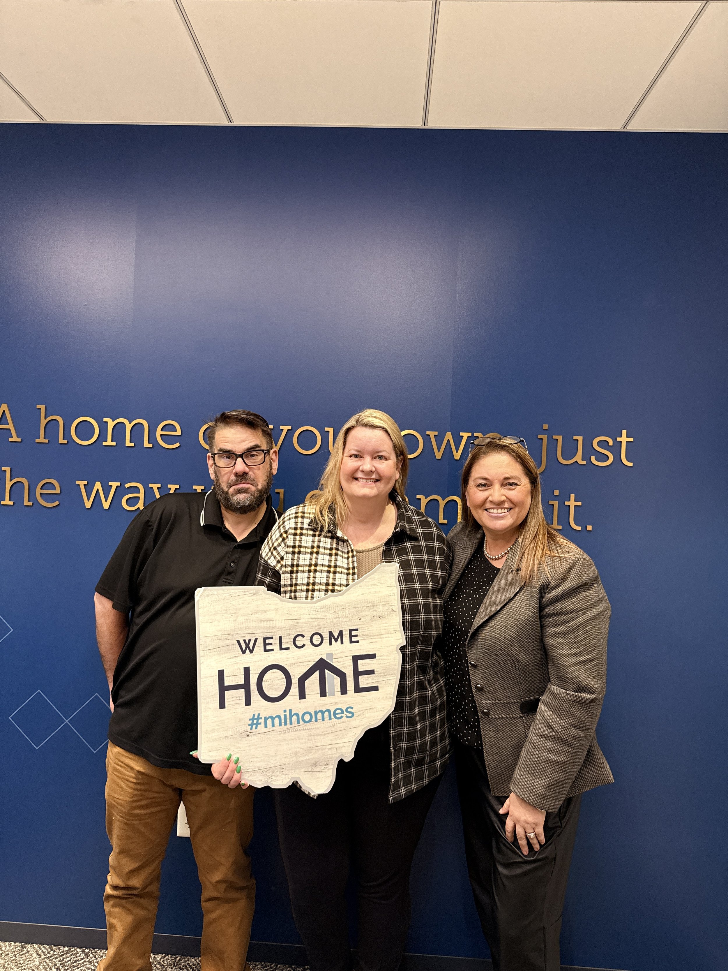 Three people standing in front of a blue wall with gold lettering. The person on the left is a man with glasses, a beard, and a black shirt. The middle person is a woman with blonde hair, wearing a plaid shirt and smiling. The person on the right is 