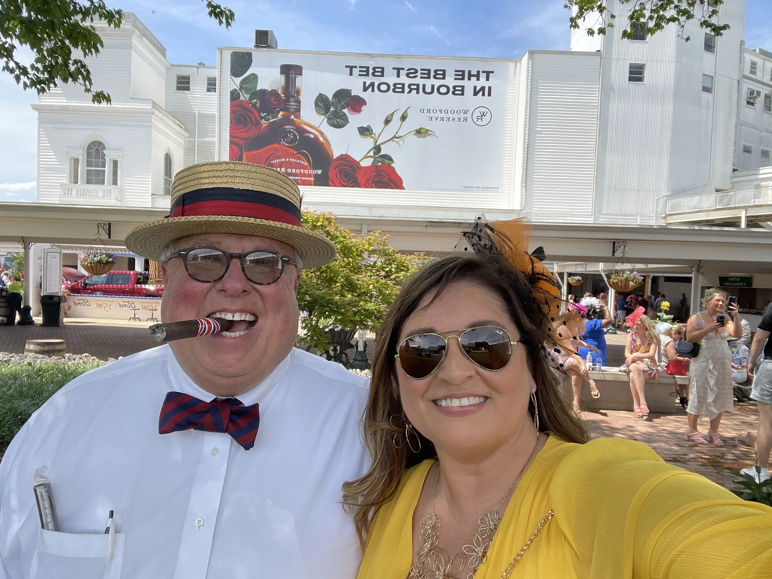 Smiling man wearing a straw hat, glasses, white shirt, and a red, white, and blue bow tie, holding a cigar, and a smiling woman wearing sunglasses, a yellow top, and jewelry, taking a selfie in front of a crowd at an outdoor event with a billboard ad