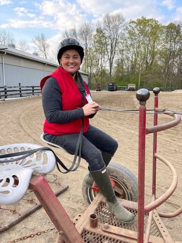 Smiling woman in riding helmet, red vest, and boots sitting on a saddleless horse riding device at an outdoor riding arena.