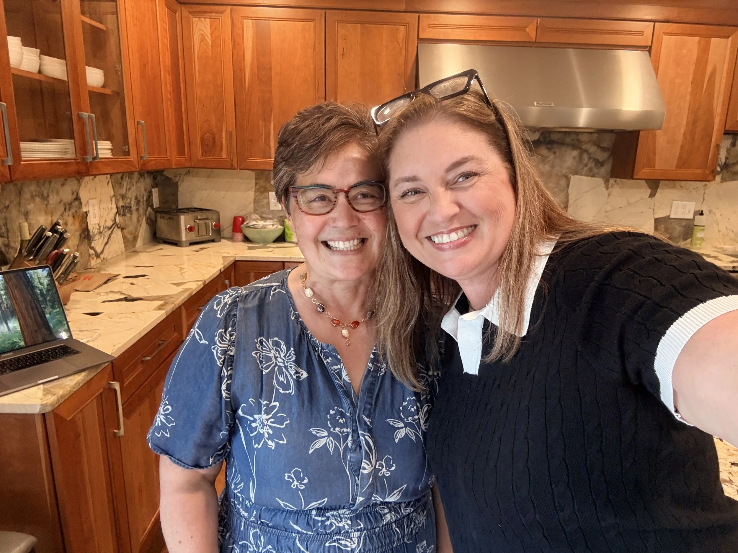 Two women are taking a selfie in a kitchen with wooden cabinets and a marble countertop. They are smiling and close together, one wearing glasses on her head and the other with glasses on her face.
