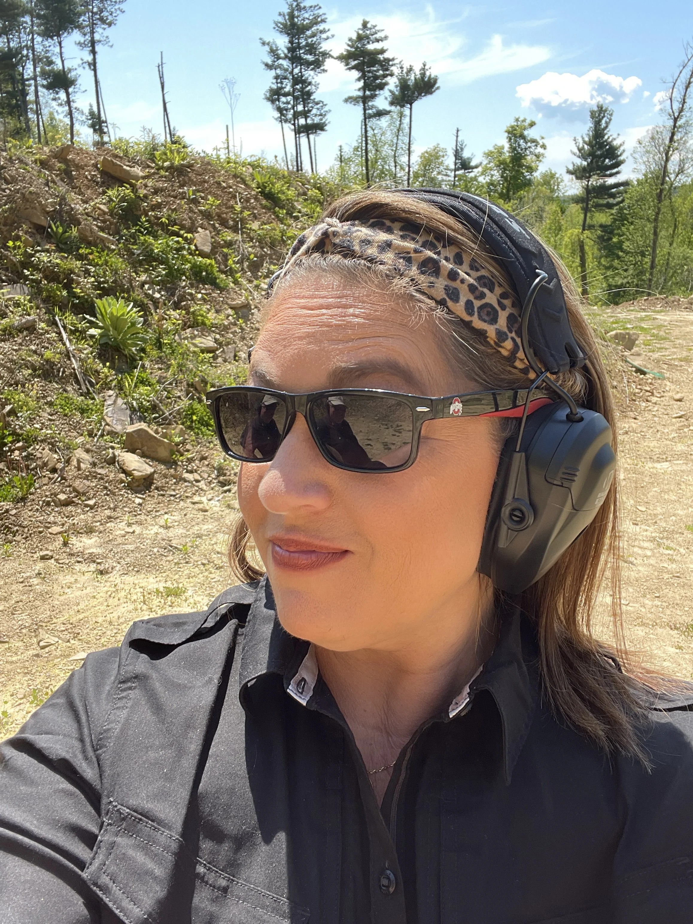 Close-up of a woman with sunglasses, a leopard print headband, black headset, and black jacket outdoors with trees and a blue sky in the background.