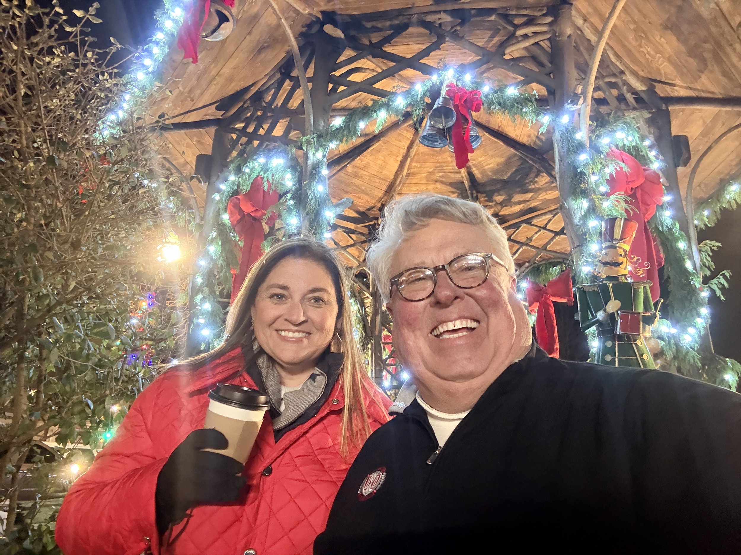 A smiling woman with long hair wearing a red jacket holds a coffee cup, standing next to a smiling man with glasses wearing a dark jacket. They are at a decorated Christmas setting with festive lights, garlands, bells, and red bows, under a wooden ro