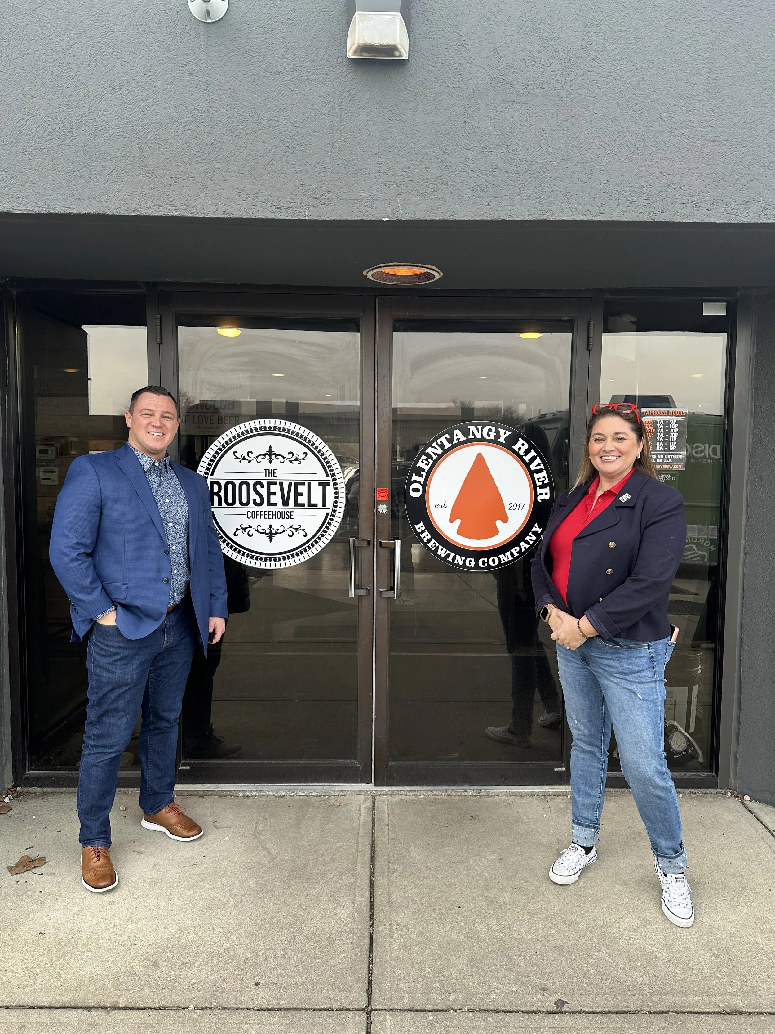 Two people standing in front of the entrance to a coffeehouse and brewing company, smiling. The man on the left is wearing a blue suit and brown shoes. The woman on the right is wearing a navy blazer with gold buttons, a red shirt, and white sneakers