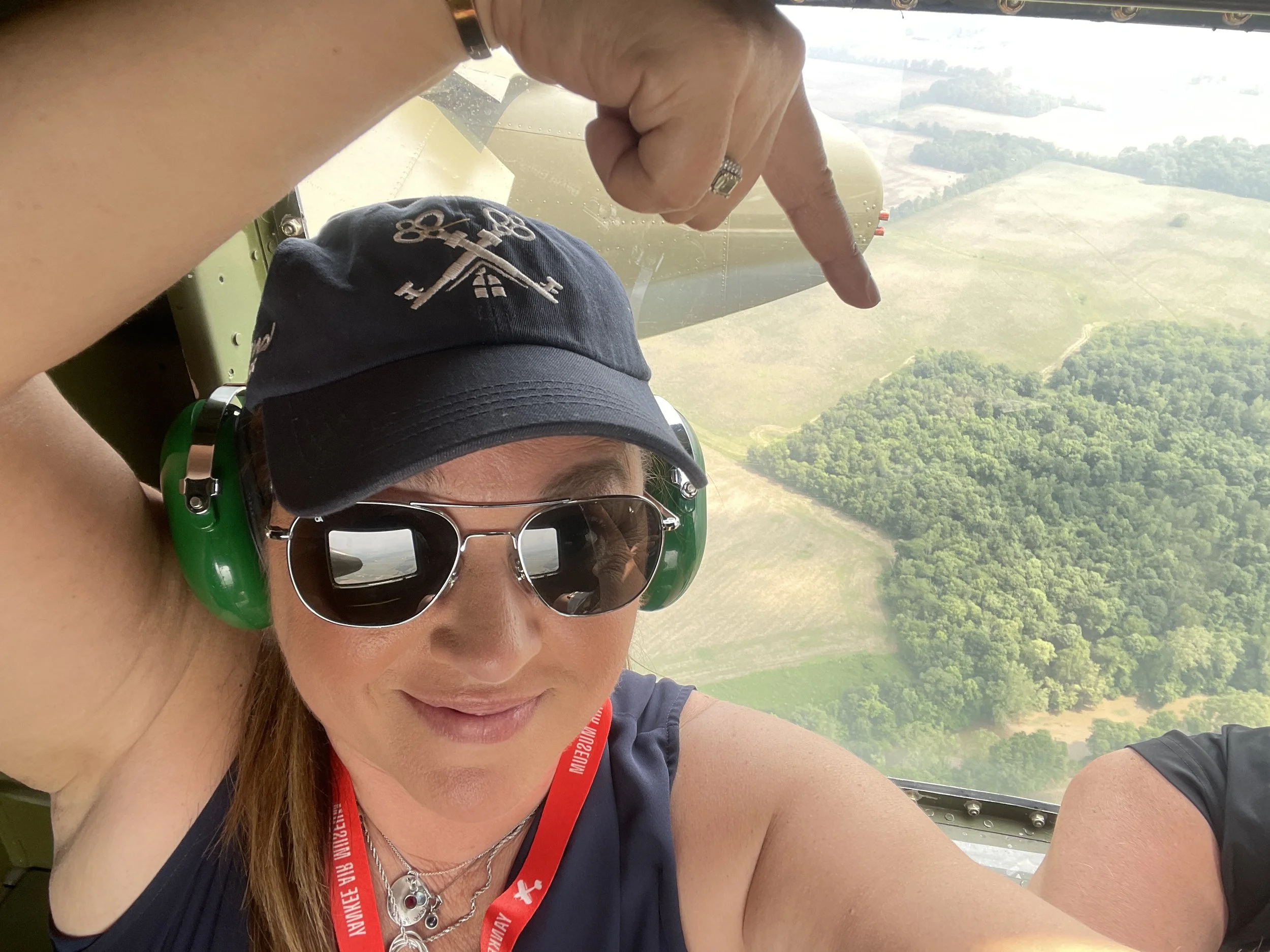 A woman with long brown hair wearing sunglasses, a black cap, and a black sleeveless top is taking a selfie while on an aerial ride in a plane. She has headphones on and a red lanyard around her neck. The plane is flying over a landscape of trees and