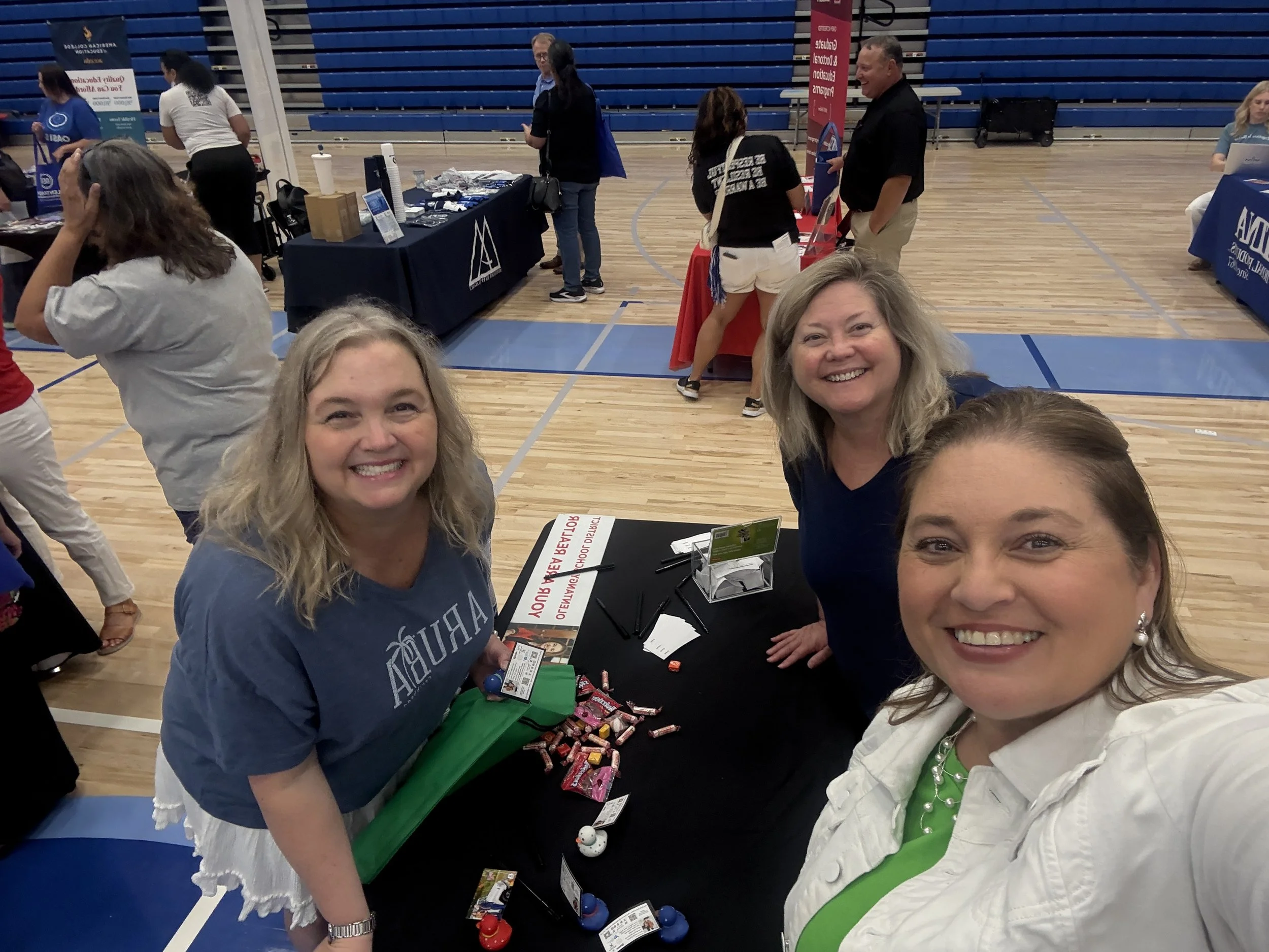People smiling and chatting at a booth in a gymnasium, with tables displaying promotional materials and candies.