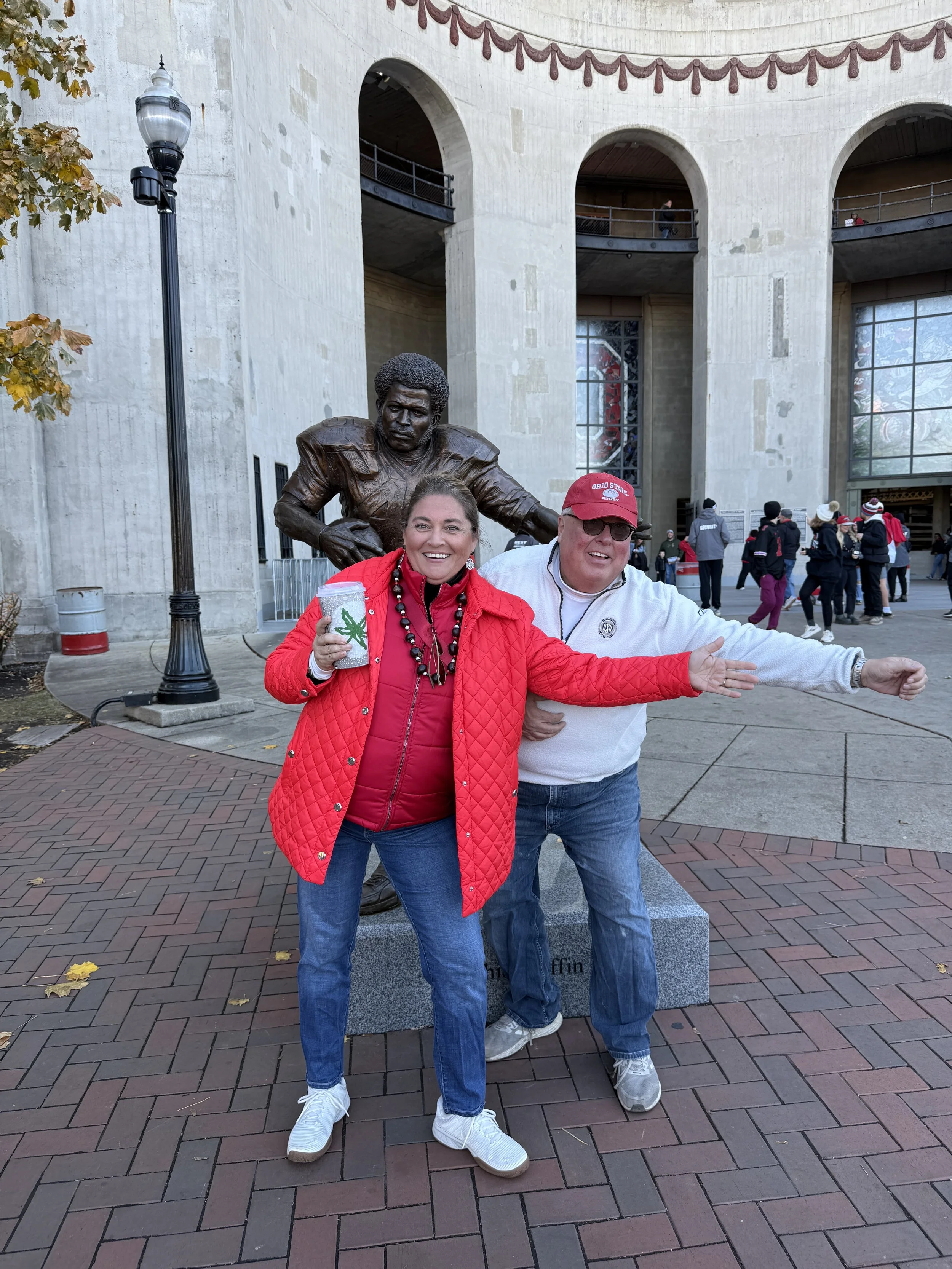 Two people, a woman in a red jacket and a man in a white jacket, stand in front of a statue of a football player on a pedestal outside an arena. The woman is smiling, holding a drink, and both are wearing casual clothes. The arena has large arched wi