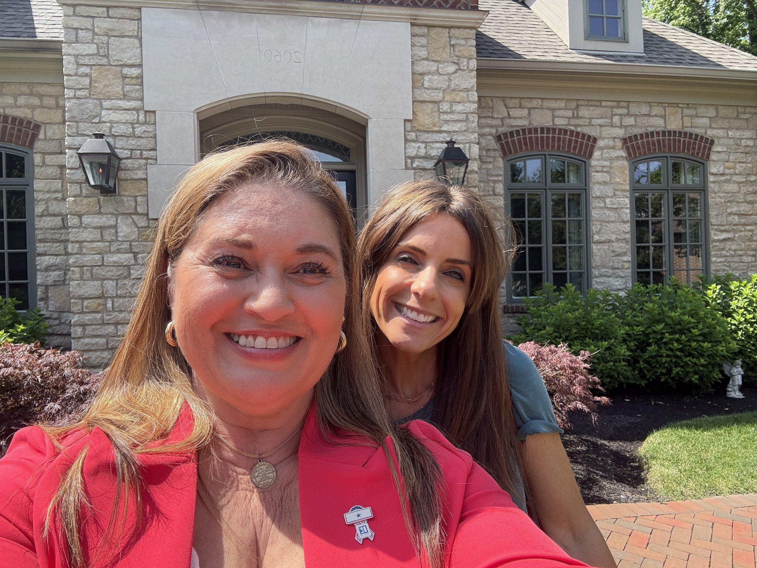 Two women smiling in front of a house with brick and stone exterior, green bushes, and a brick walkway.