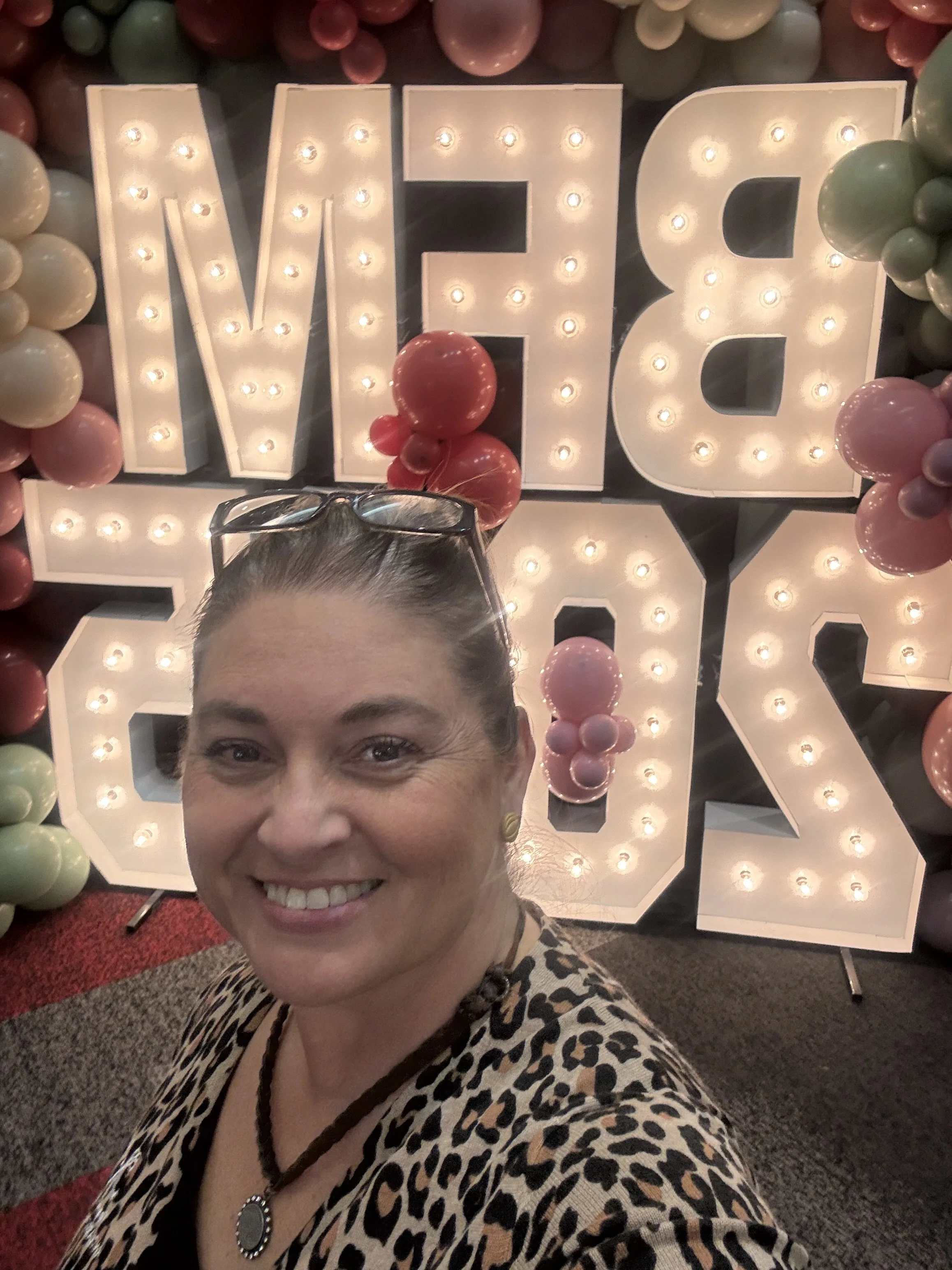 A woman smiling in front of a decorative sign spelling 'LOVE' with illuminated letters, surrounded by colorful balloons.
