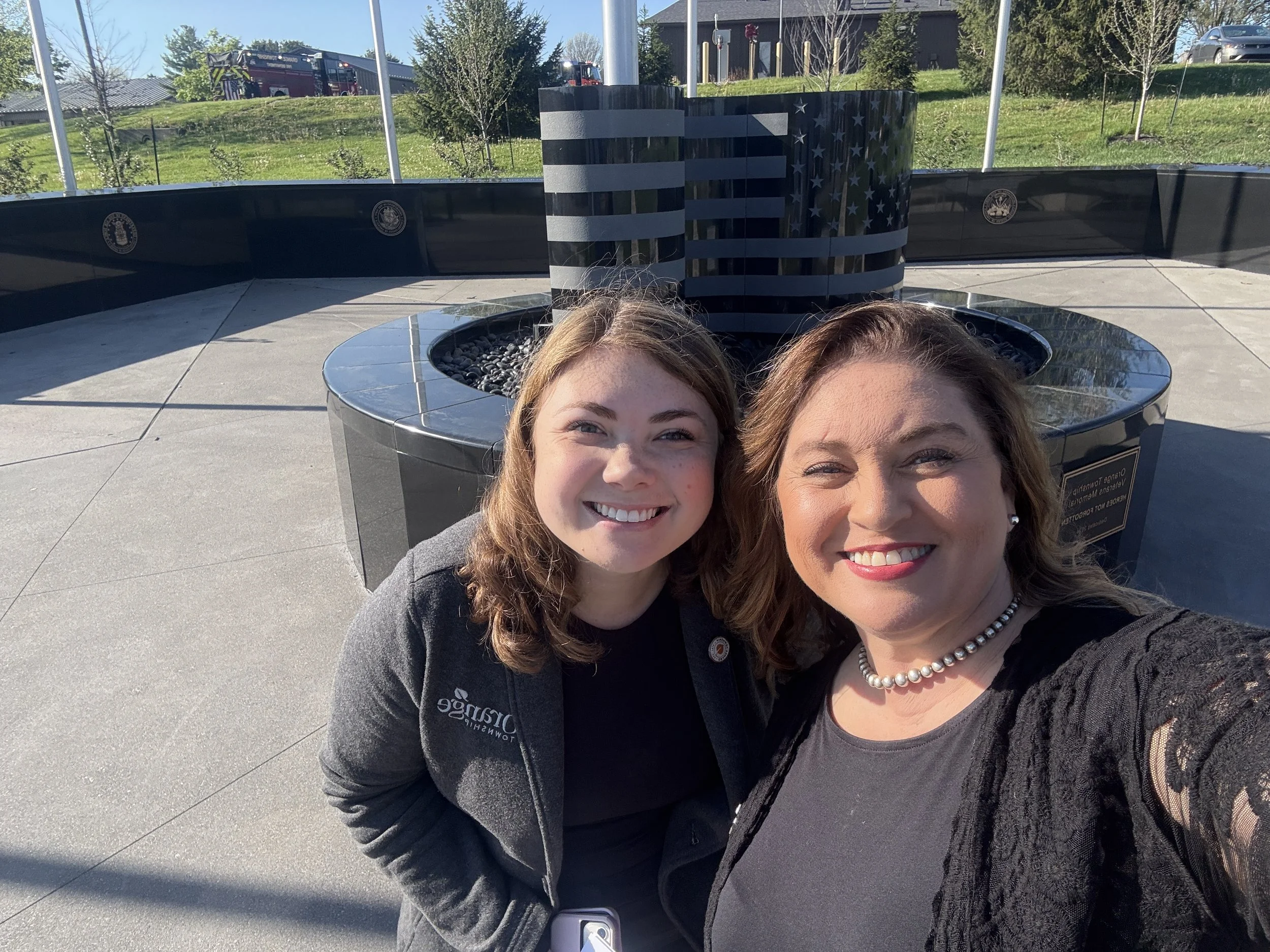 Two women taking a selfie outdoors, with a black memorial monument featuring the American flag's black and white stripes and stars in the background.