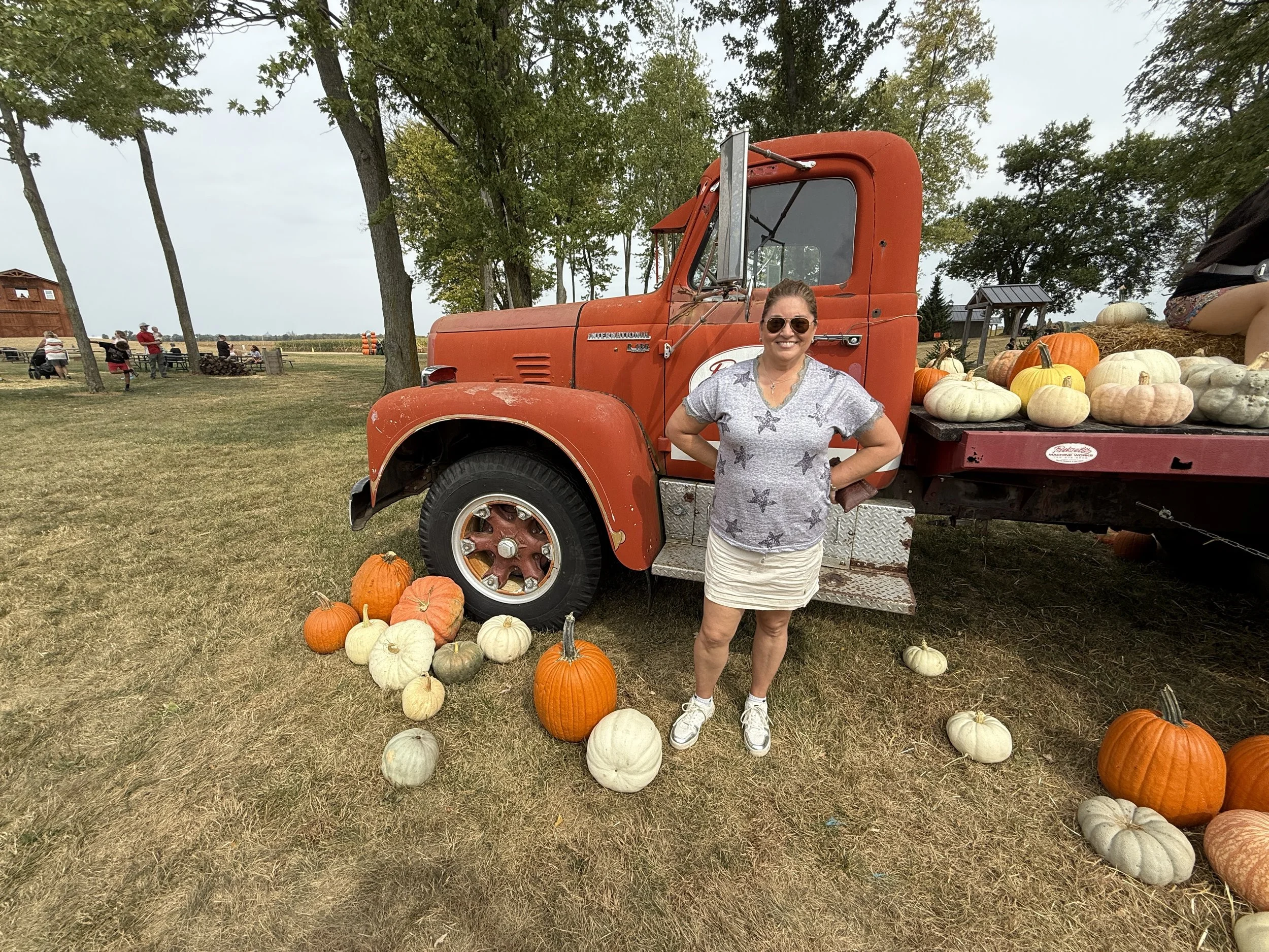 A woman wearing sunglasses and a star-patterned shirt stands in front of a red vintage truck decorated with pumpkins at a fall pumpkin patch, with trees and other visitors in the background.