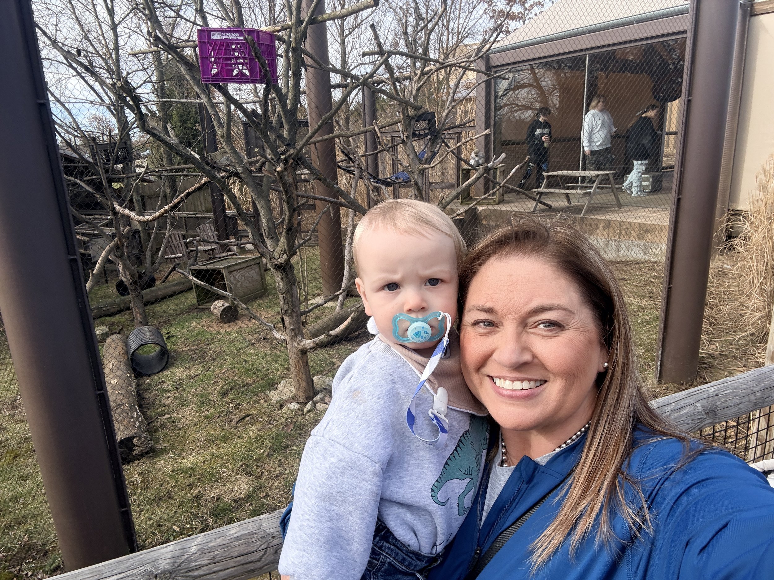 A woman and a young child taking a selfie outside in front of a fenced area with trees and a small building, with three people standing in the background.