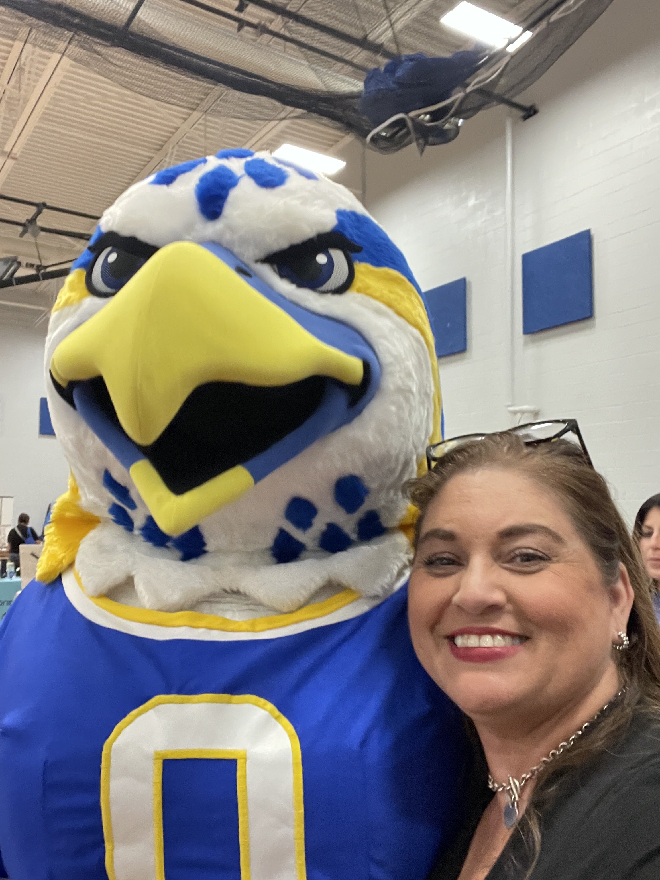 Close-up of a person smiling with a large blue, white, and yellow mascot costume resembling a bird, possibly an eagle or a hawk, in an indoor setting.