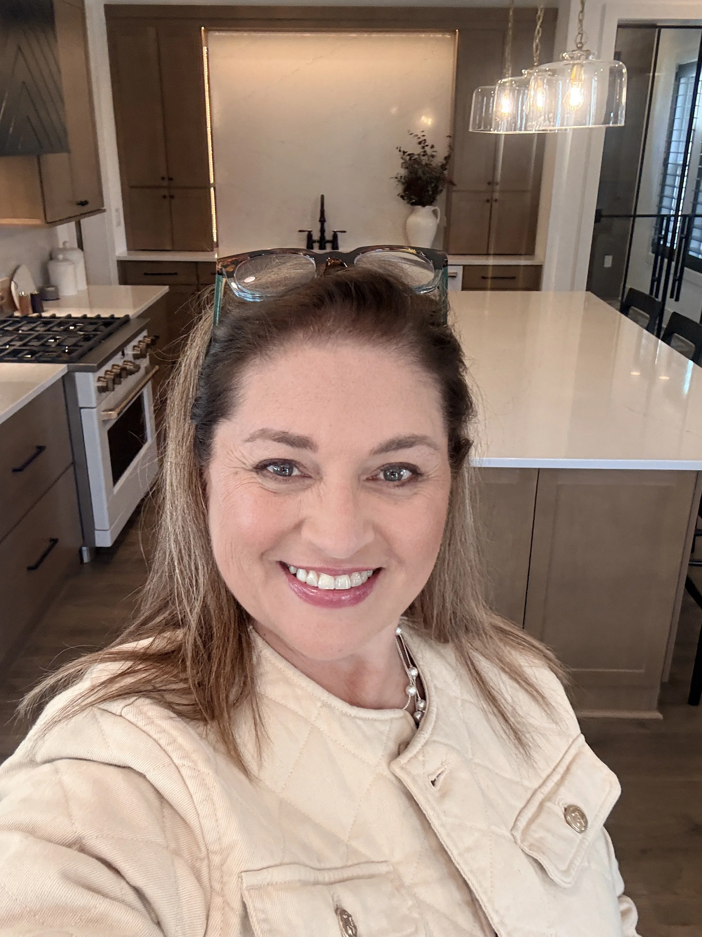 A smiling woman with glasses on her head, wearing a beige jacket and pearl necklace, taking a selfie in a modern kitchen with a large island, wooden cabinets, and a hanging glass light fixture.
