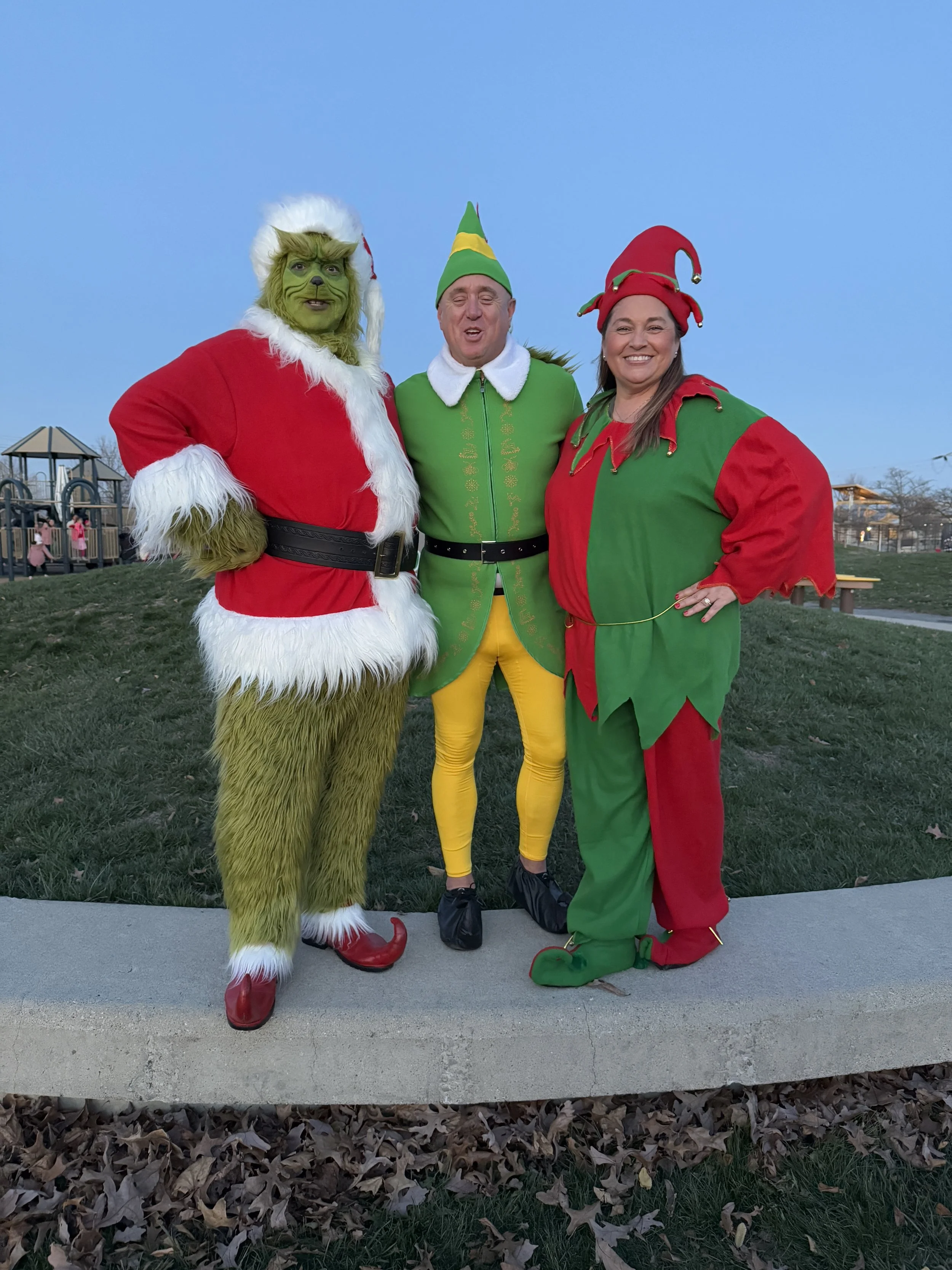 Three people dressed as Christmas characters, including a person in a Grinch costume, a person dressed as an elf, and a person dressed as an elf with a jester hat, standing outdoors on grass with a playground and trees in the background.