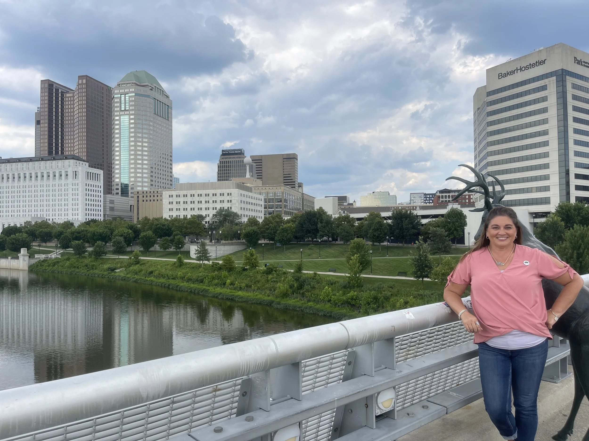 City skyline with tall buildings, a river, and a park with trees in the foreground, taken from a bridge. A woman in a pink blouse stands by the bridge railing, smiling for the photo.
