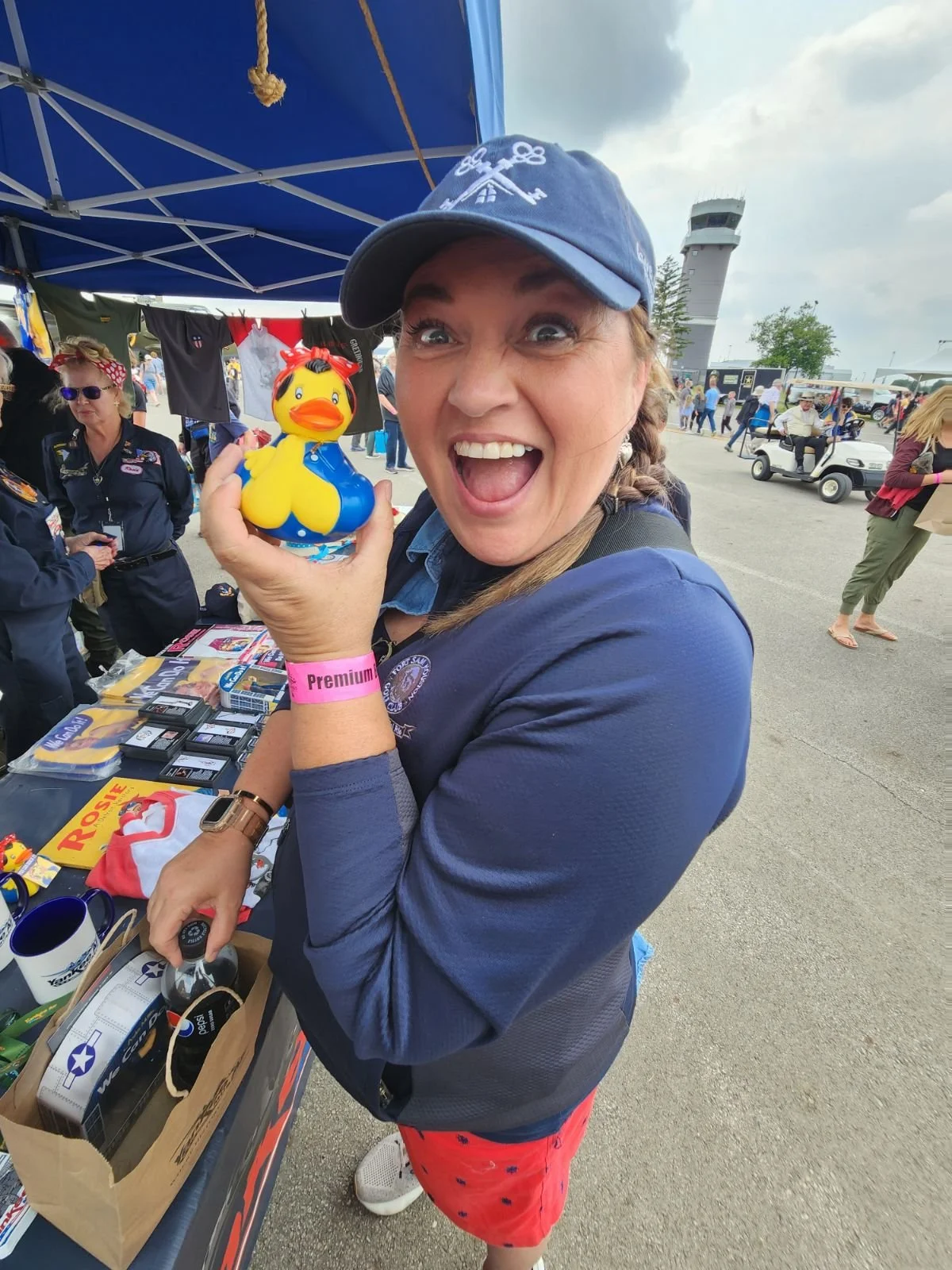A woman with braided hair, wearing a navy jacket and a blue cap, is smiling widely and holding a yellow rubber duck with a blue sweater. She is at an outdoor event with booths, people, and a control tower in the background.