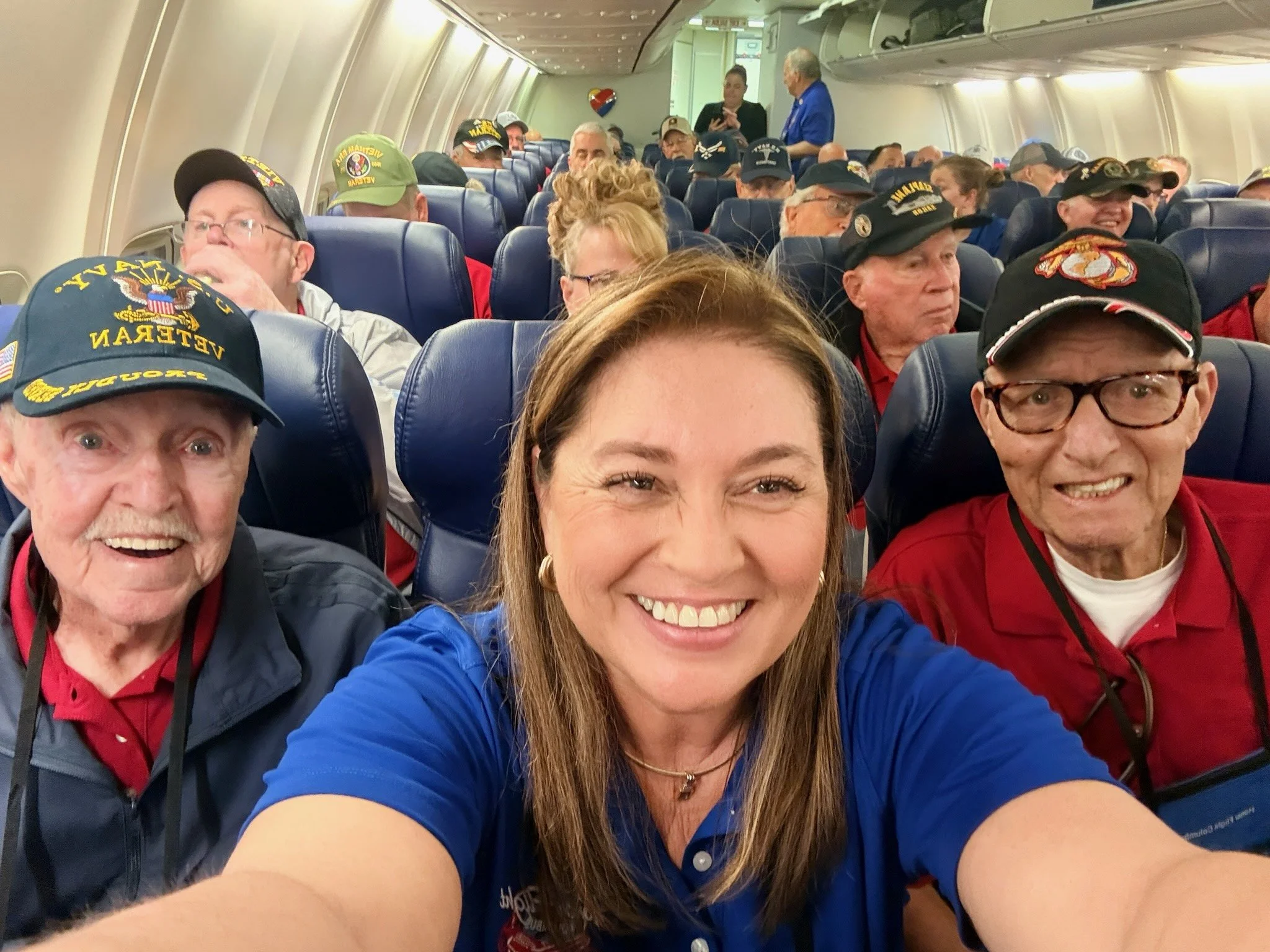 Interior of an airplane filled with mostly elderly passengers, many wearing military veterans hats, taking a selfie, smiling, with a flight attendant in the background.