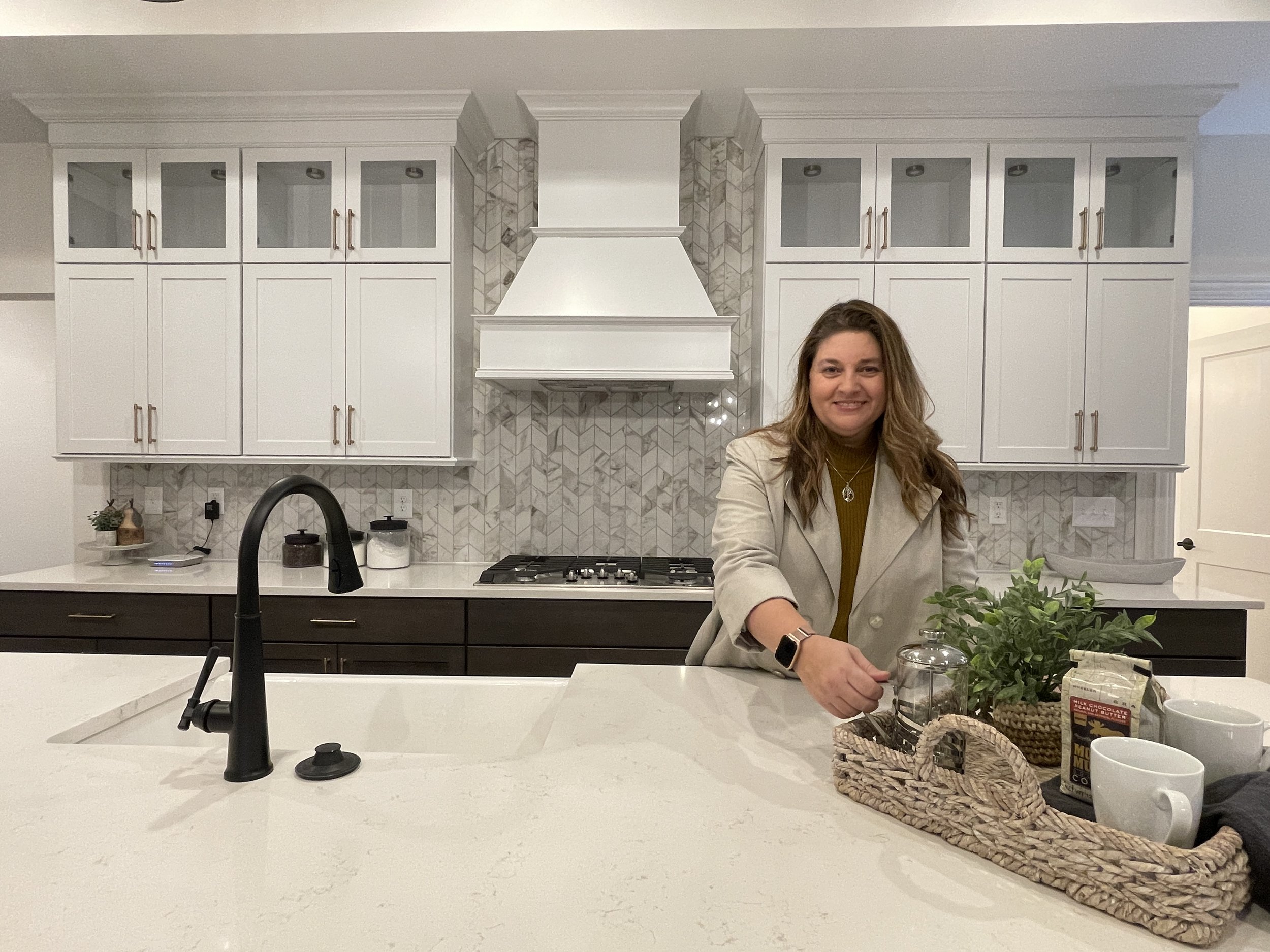 A woman smiling at a kitchen counter with a plant, basket, and coffee mugs, in front of white cabinets and a decorative backsplash.