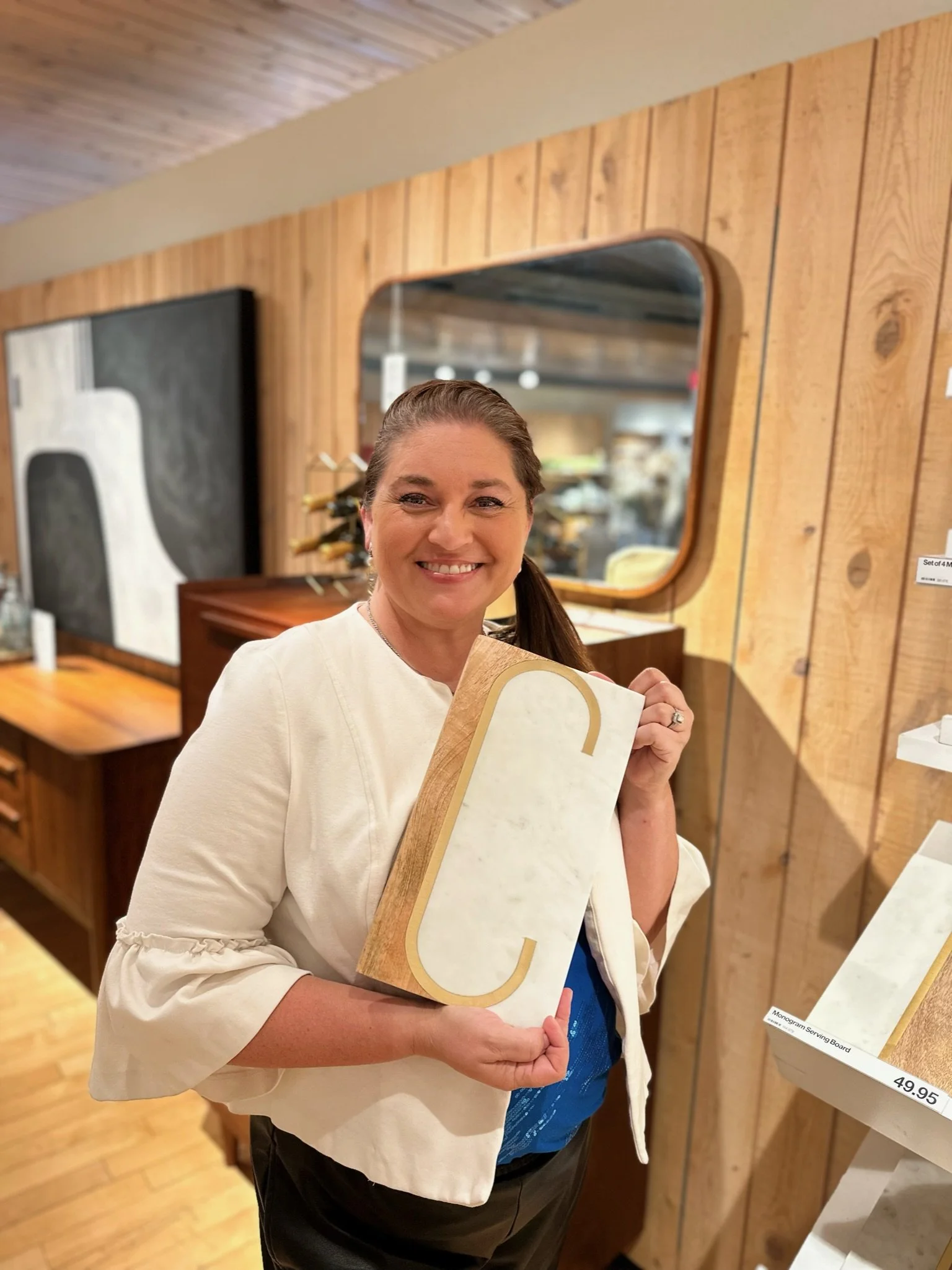A smiling woman with brown hair pulled back, holding a large wooden and white marble serving board with a gold border, standing in a store with wooden walls and a mirror in the background.